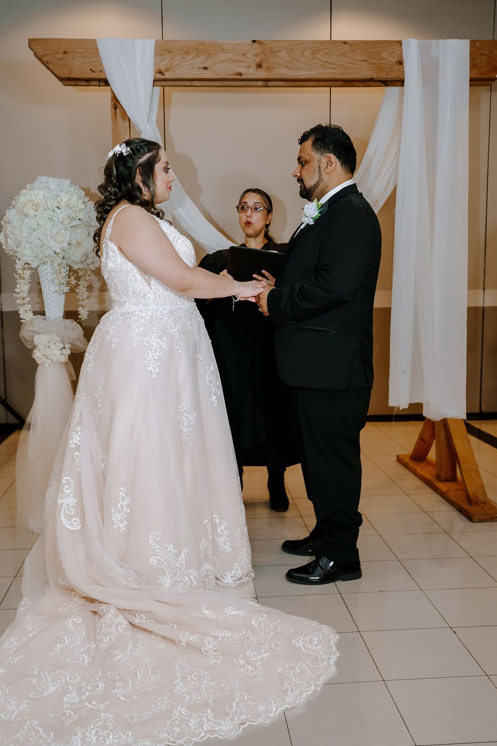 Elegant wedding scene showing a bride in a white lace gown dancing with someone in a black suit on a tiled floor.