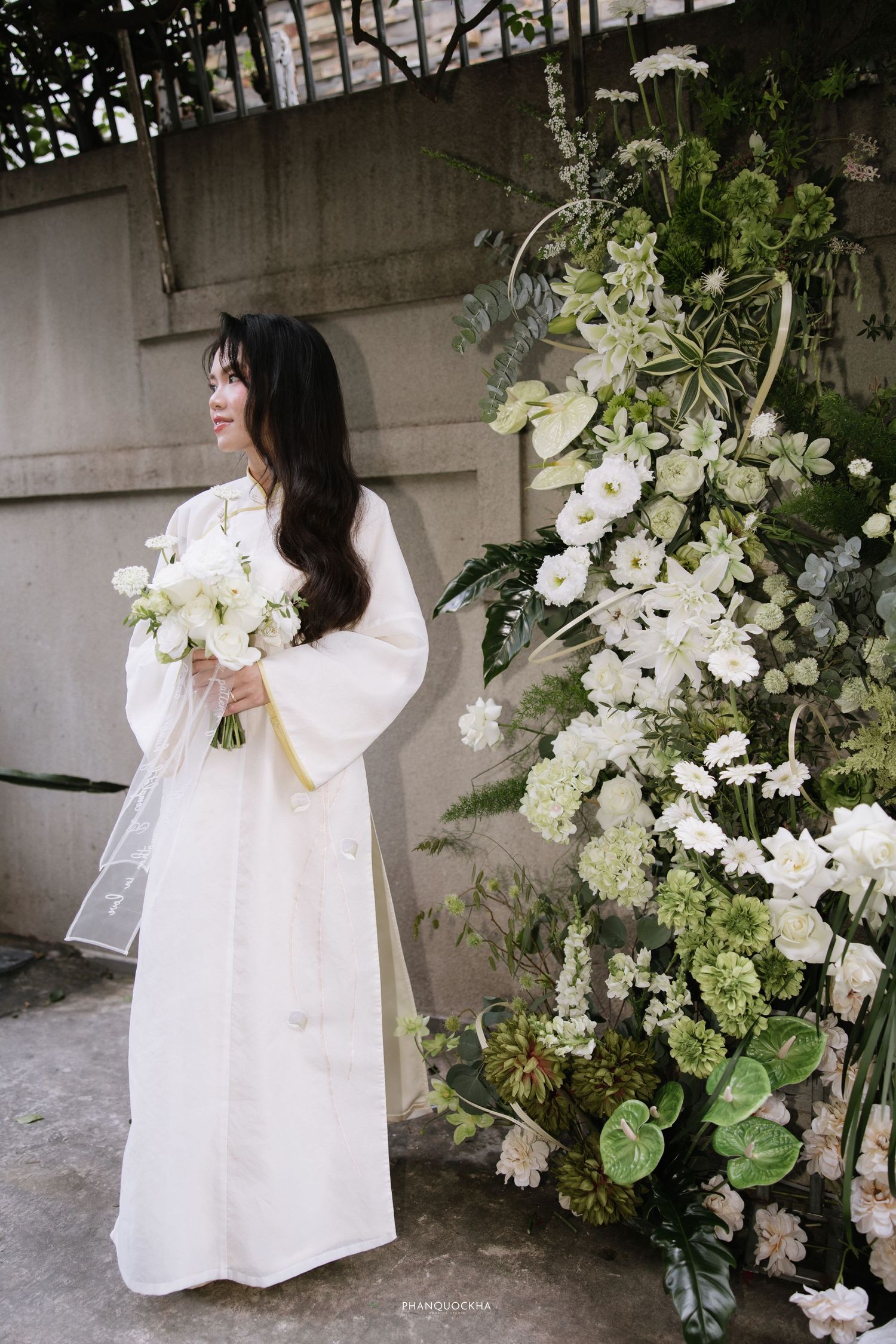 Bride in white linen dress holds bouquet beside tall green and white floral arrangement outdoors.