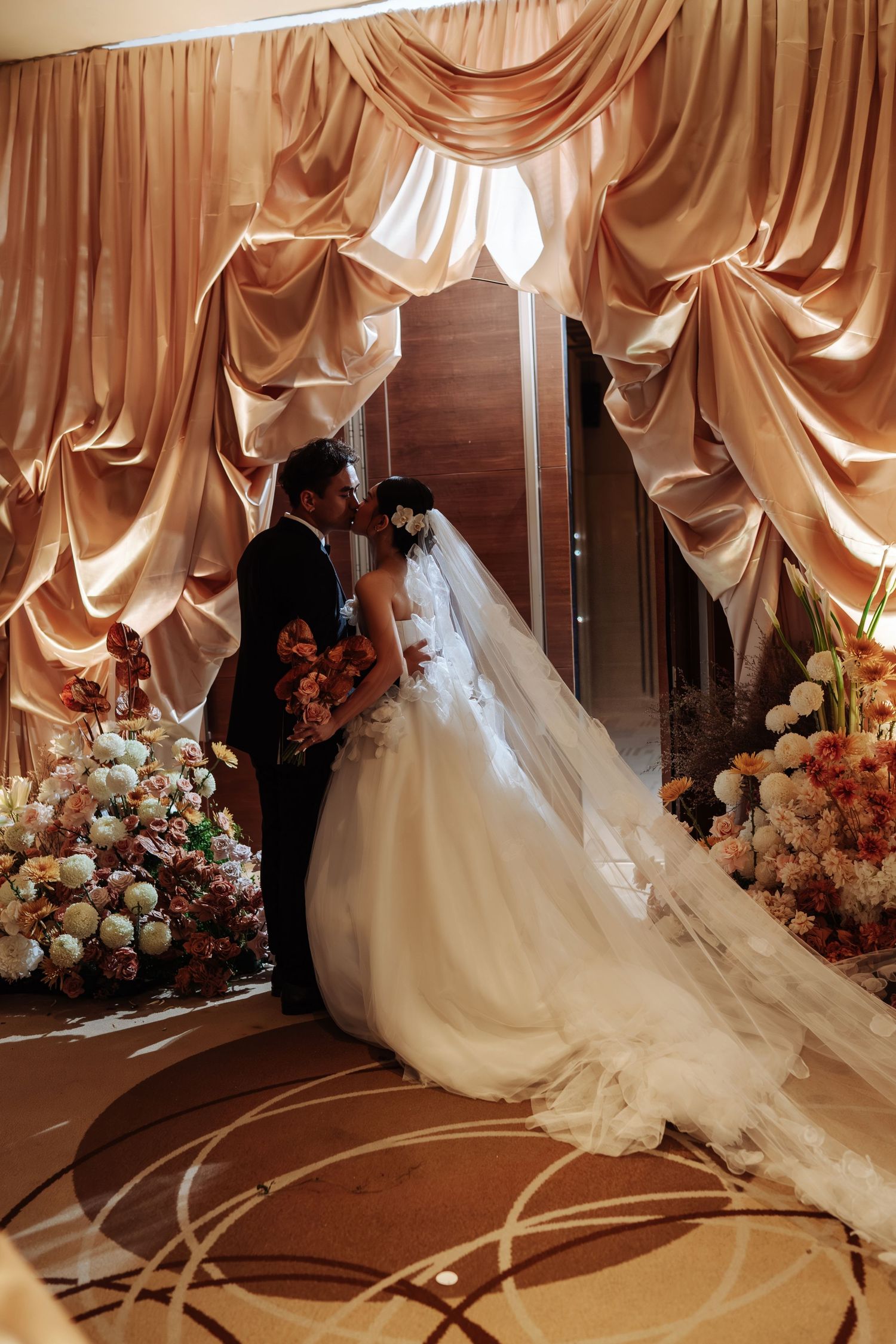 Bride and groom share a romantic kiss under elegant golden draped curtains surrounded by lush floral arrangements.