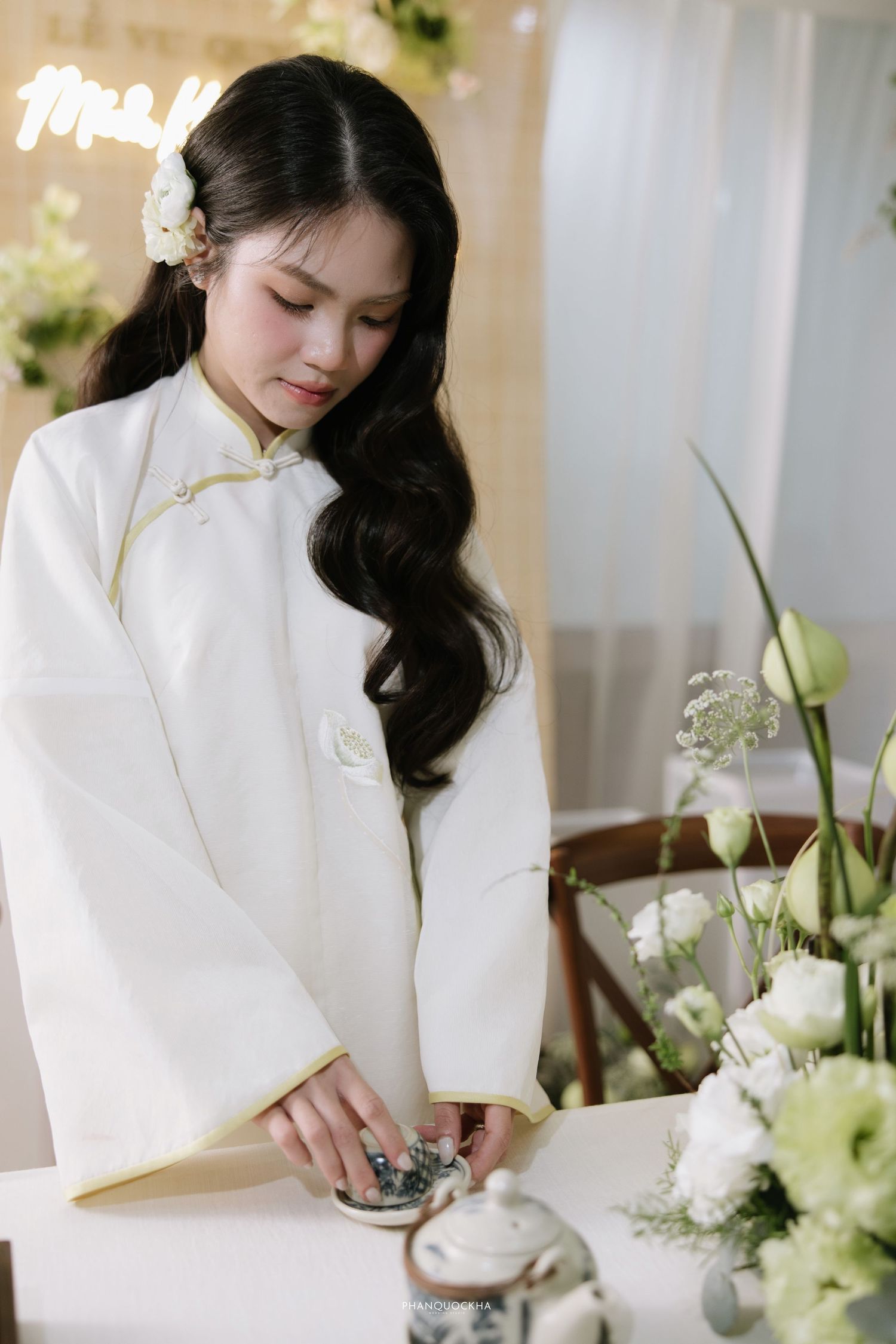 Beautiful bride in white ao dai with gold trim arranges items on table with white floral decor.