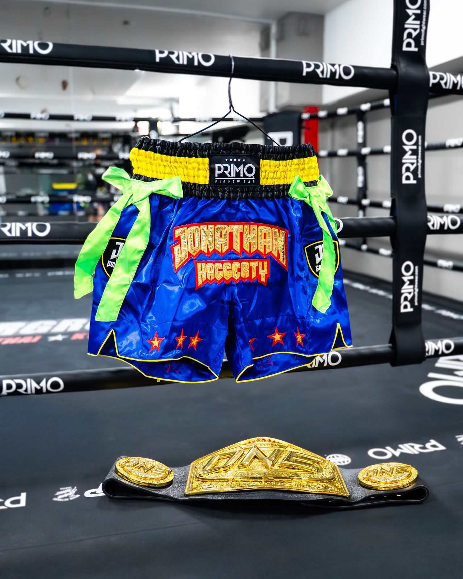 Vibrant blue and neon green Muay Thai boxing shorts displayed with championship belt in a boxing ring setting.