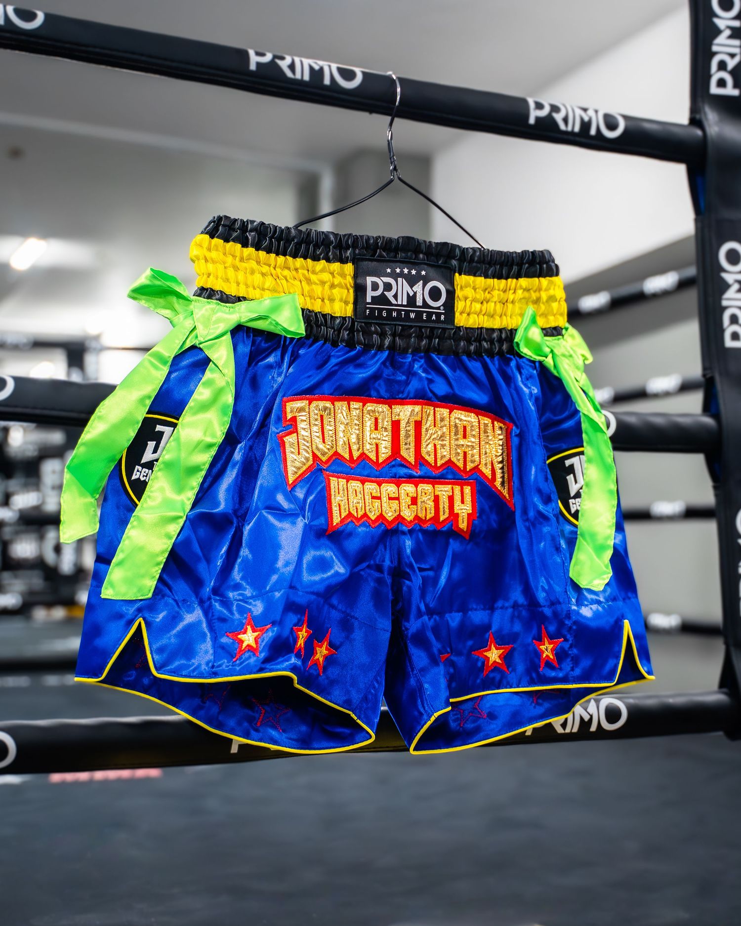 Custom blue Muay Thai shorts with yellow waistband and neon green side ties hanging in a boxing gym.