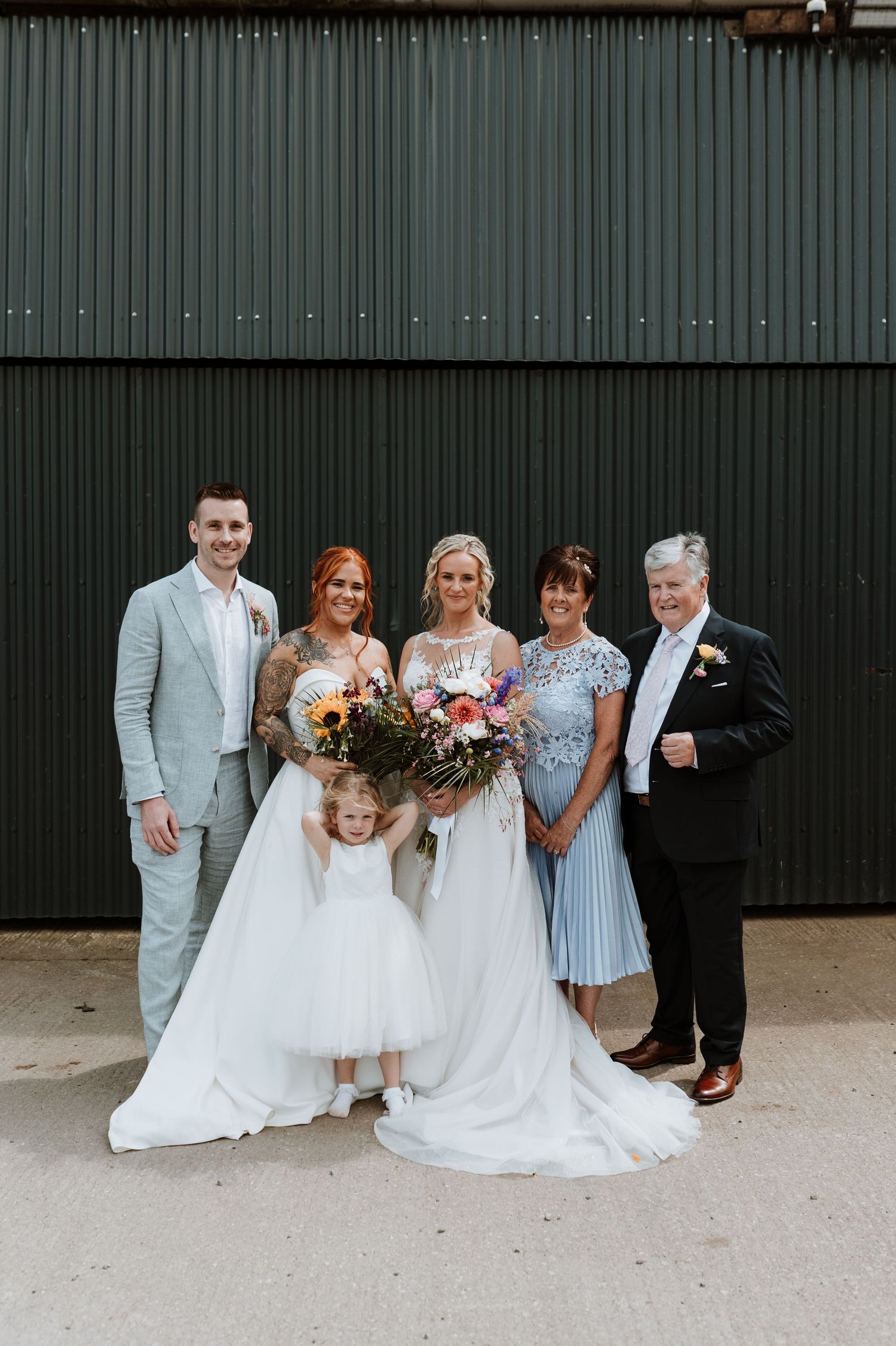 Wedding party poses together wearing elegant white dresses and light gray suits in front of dark metal wall at Stanford Farm