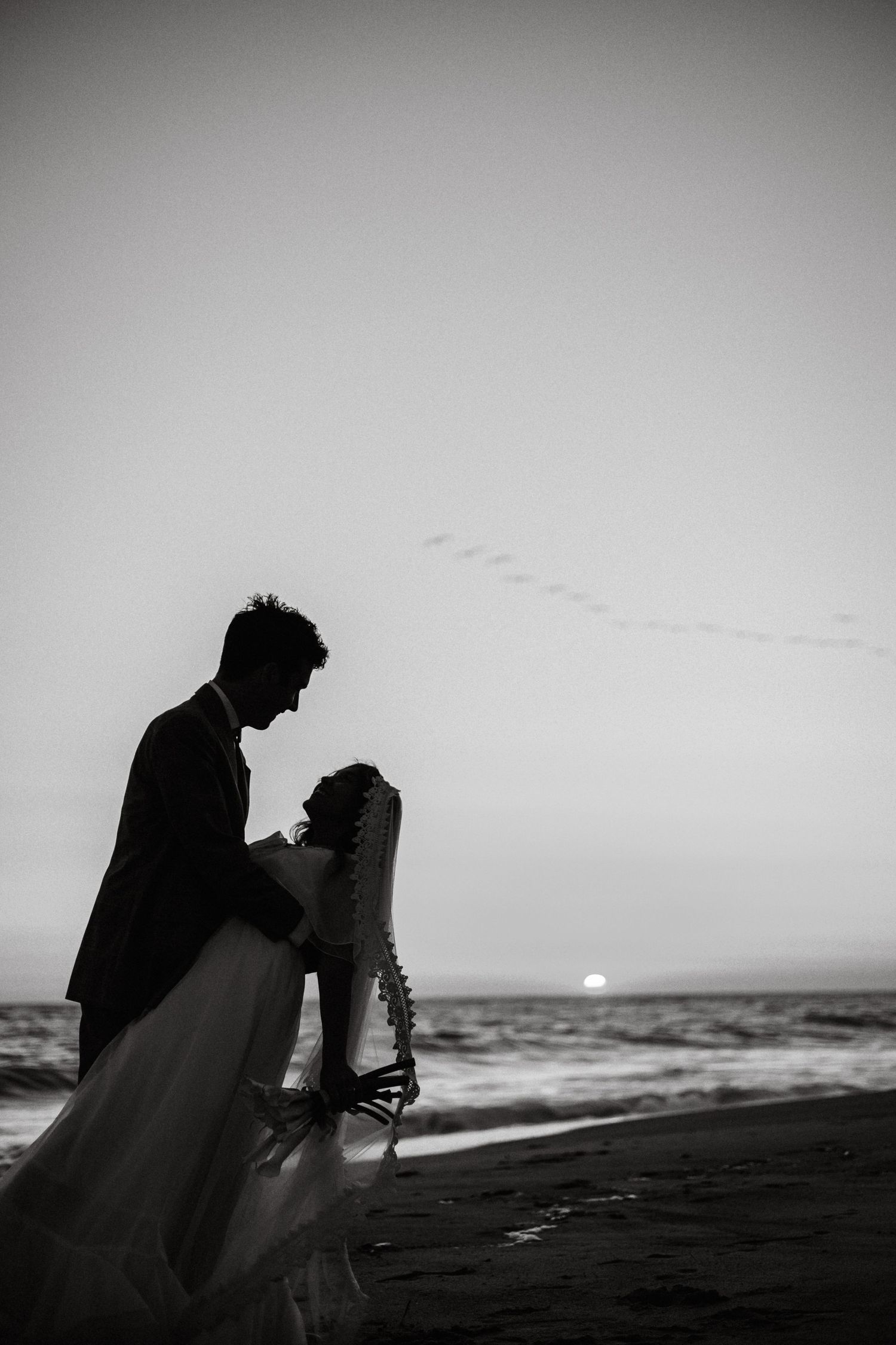 A black and white photo of a couple in wedding clothes embracing one another on the beach with brids flying and a sunset in the background.