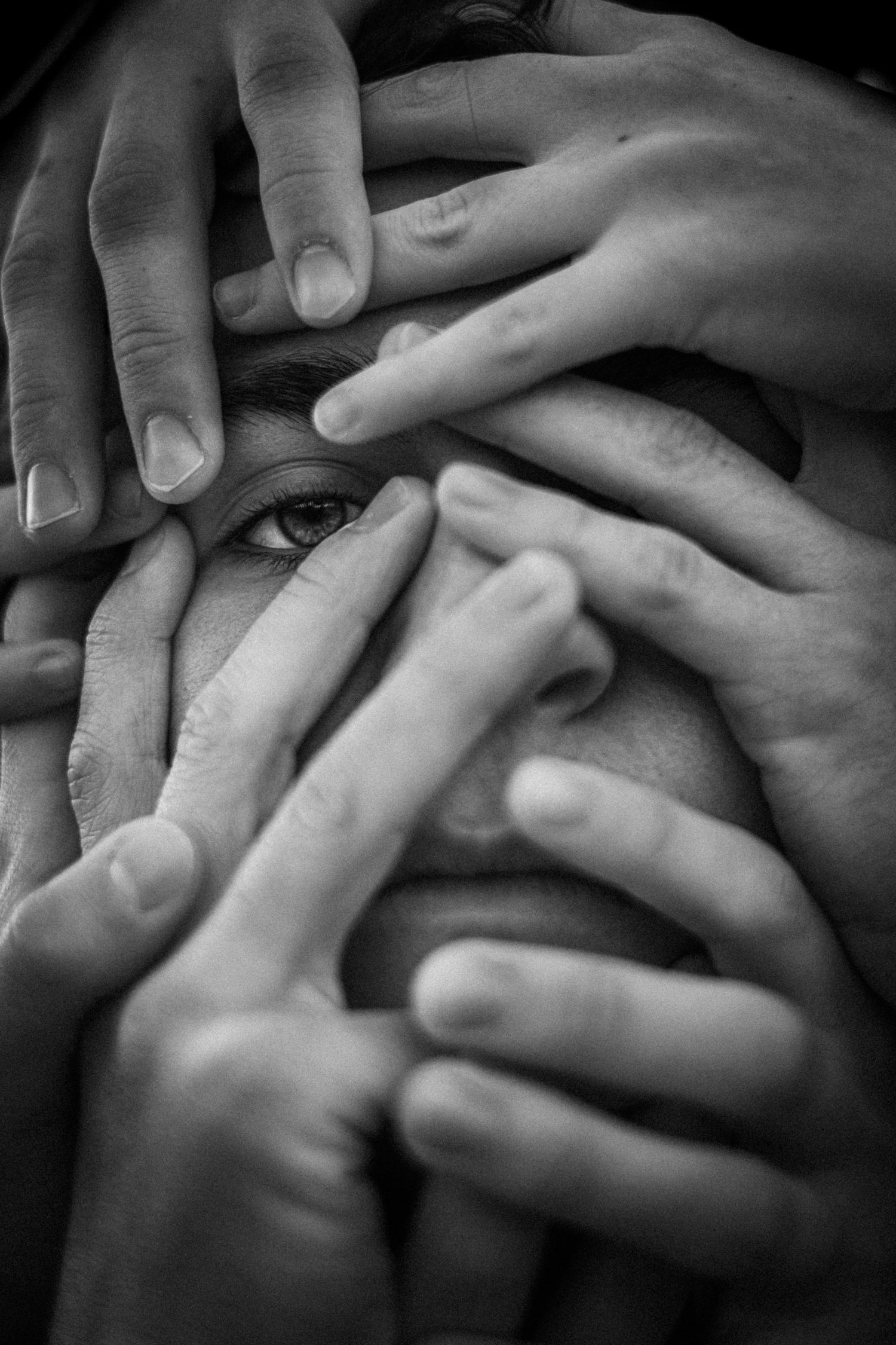 Numerous hands cover a face, with a single eye and a portion of the mouth visible through the fingers.