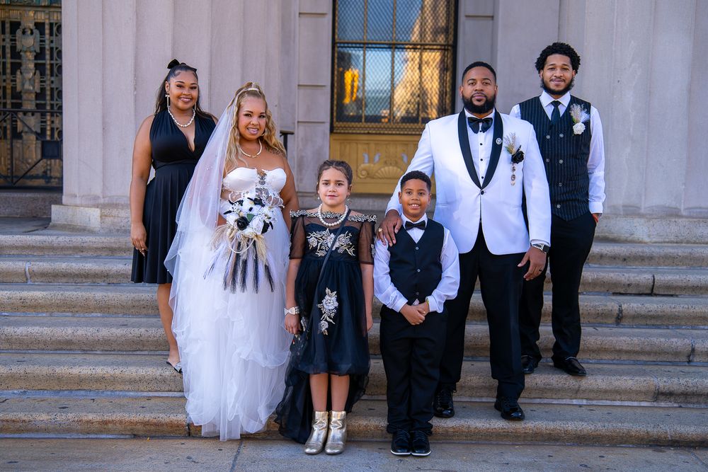 Wedding party poses on grand stone steps outside historic building in formal attire with matching white and navy outfits.