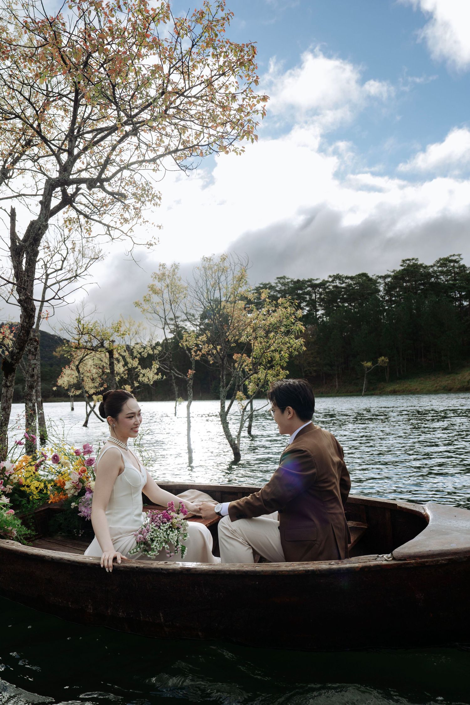 Bride and groom hold hands in flower-filled wooden boat on serene lake surrounded by autumn trees.