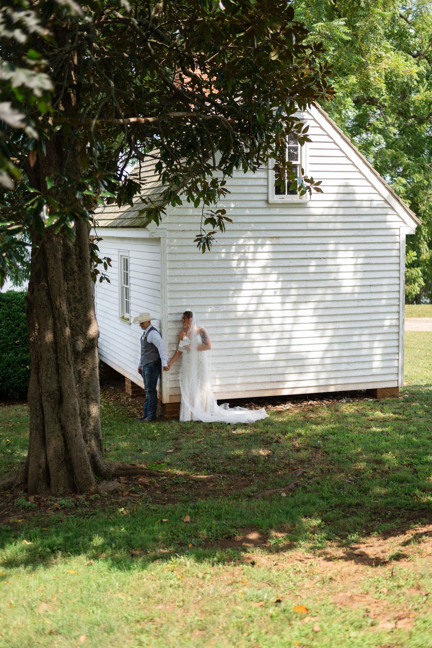 A white wooden outbuilding with siding stands in a shaded yard beneath a large tree on a sunny summer day.