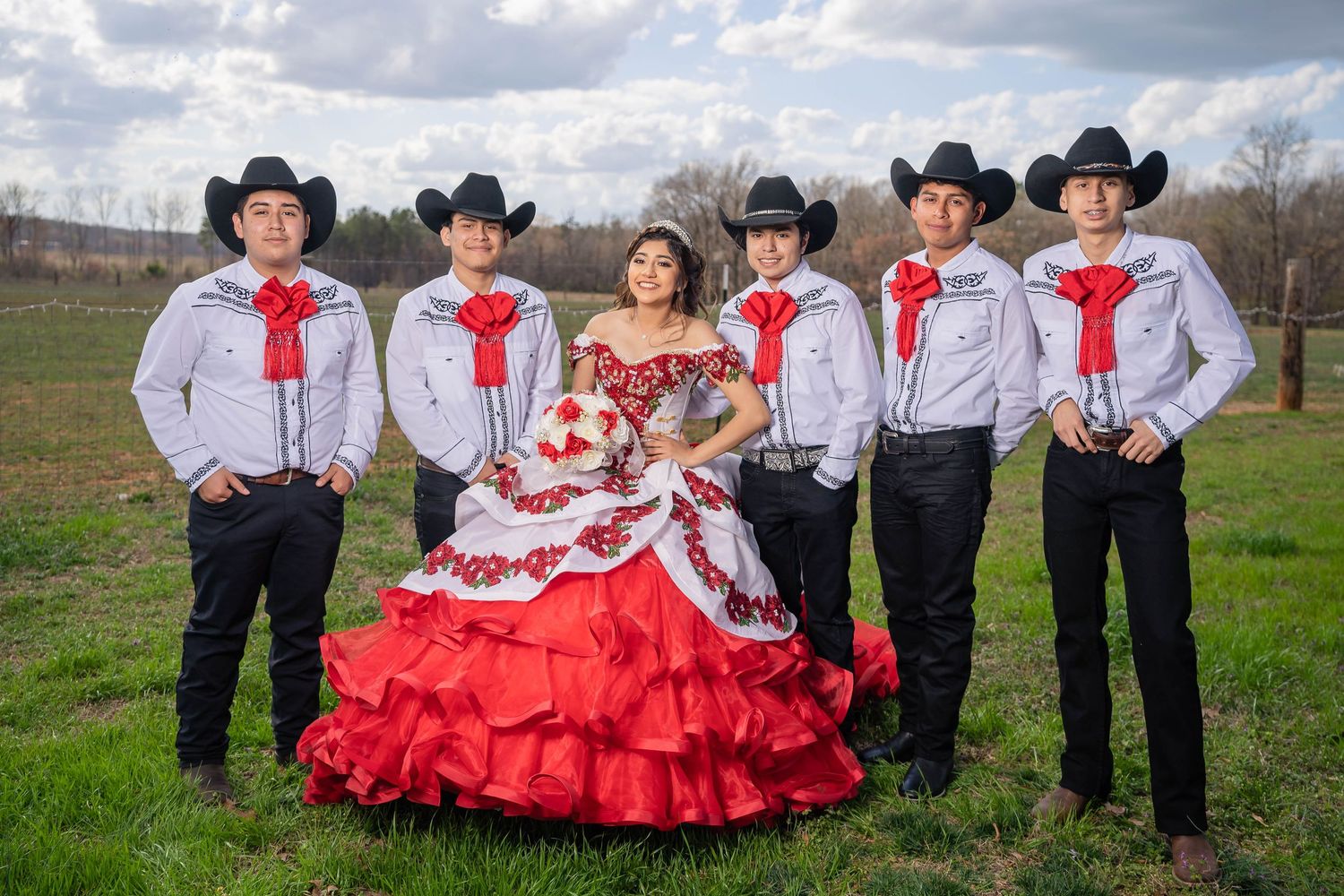 Western dance performers in black cowboy hats and red and white outfits pose together on a grassy field.