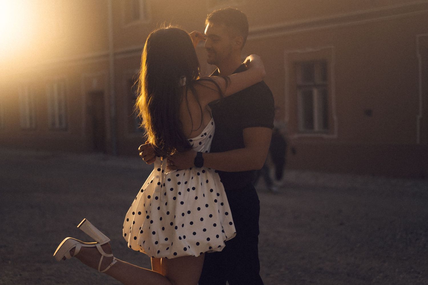 A person in a polka dot dress walks through a sunlit cobblestone street with historic buildings in the background.