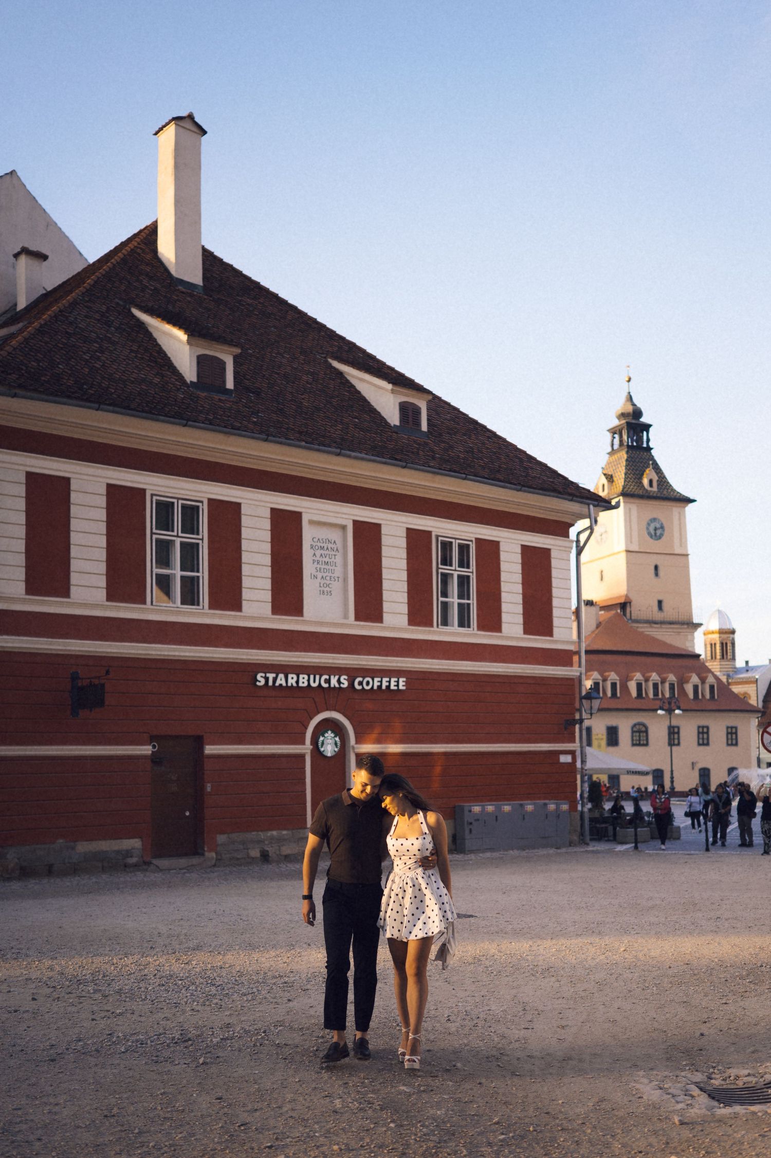 A couple walks past a Starbucks Coffee shop in a historic European town square with a church tower visible in the background.