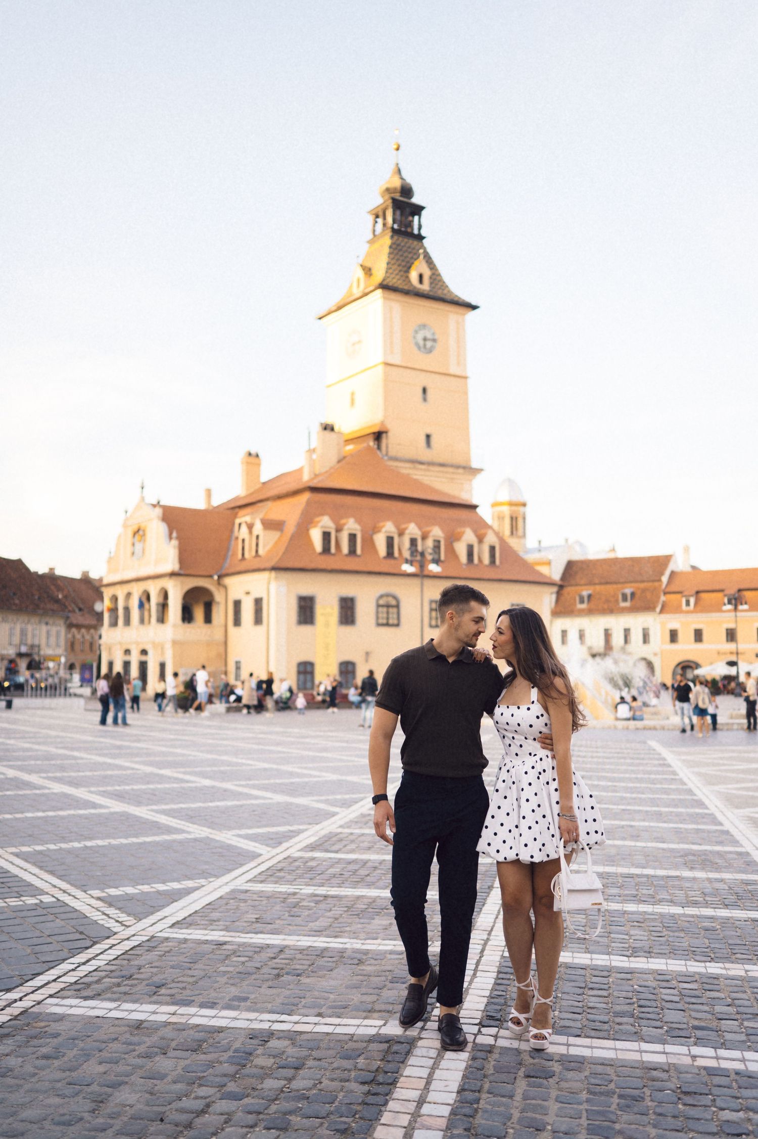 A couple walks across a historic European town square with a grand church tower and traditional architecture in the background.