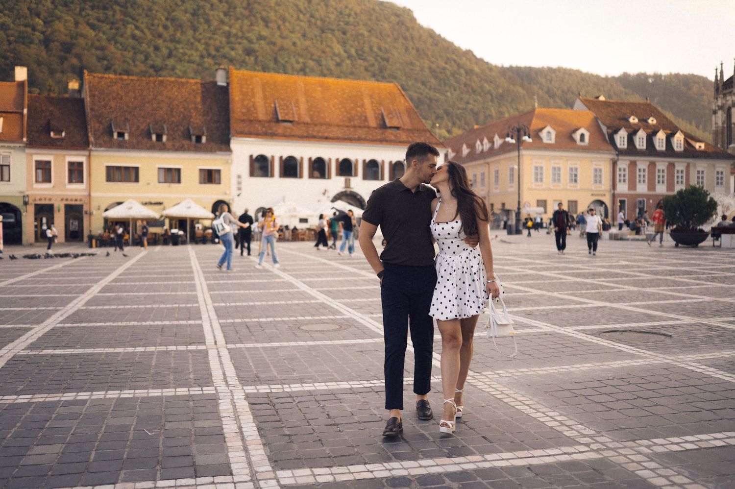 People walk across a large stone paved plaza in front of historic European buildings on a sunny day.