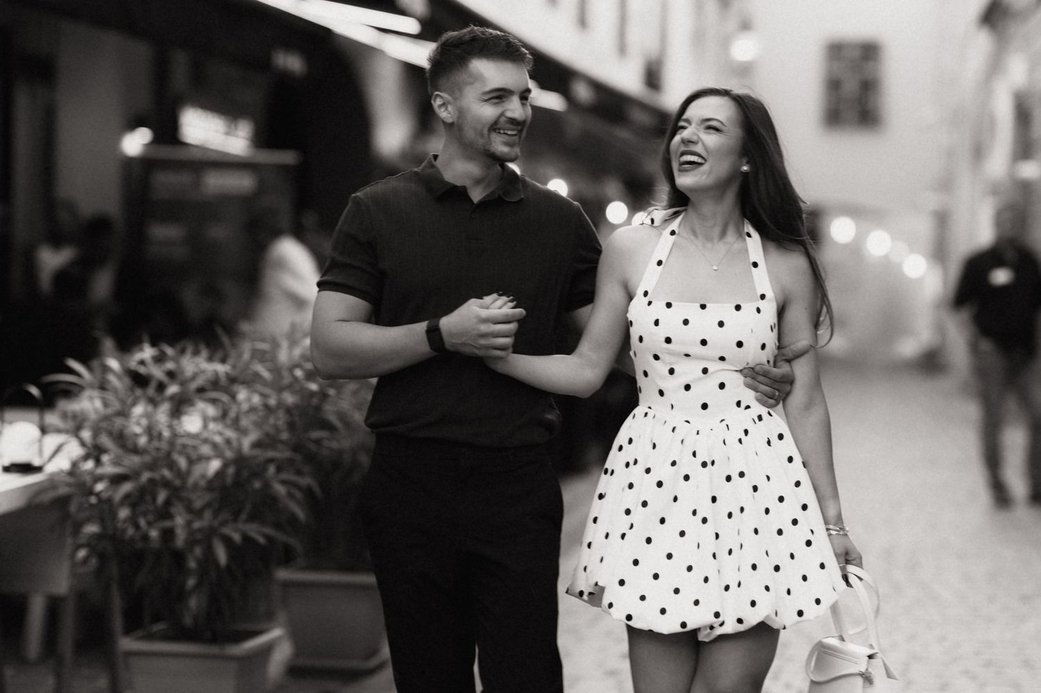 A couple laughs together while walking down a city street, with the woman wearing a polka dot dress.