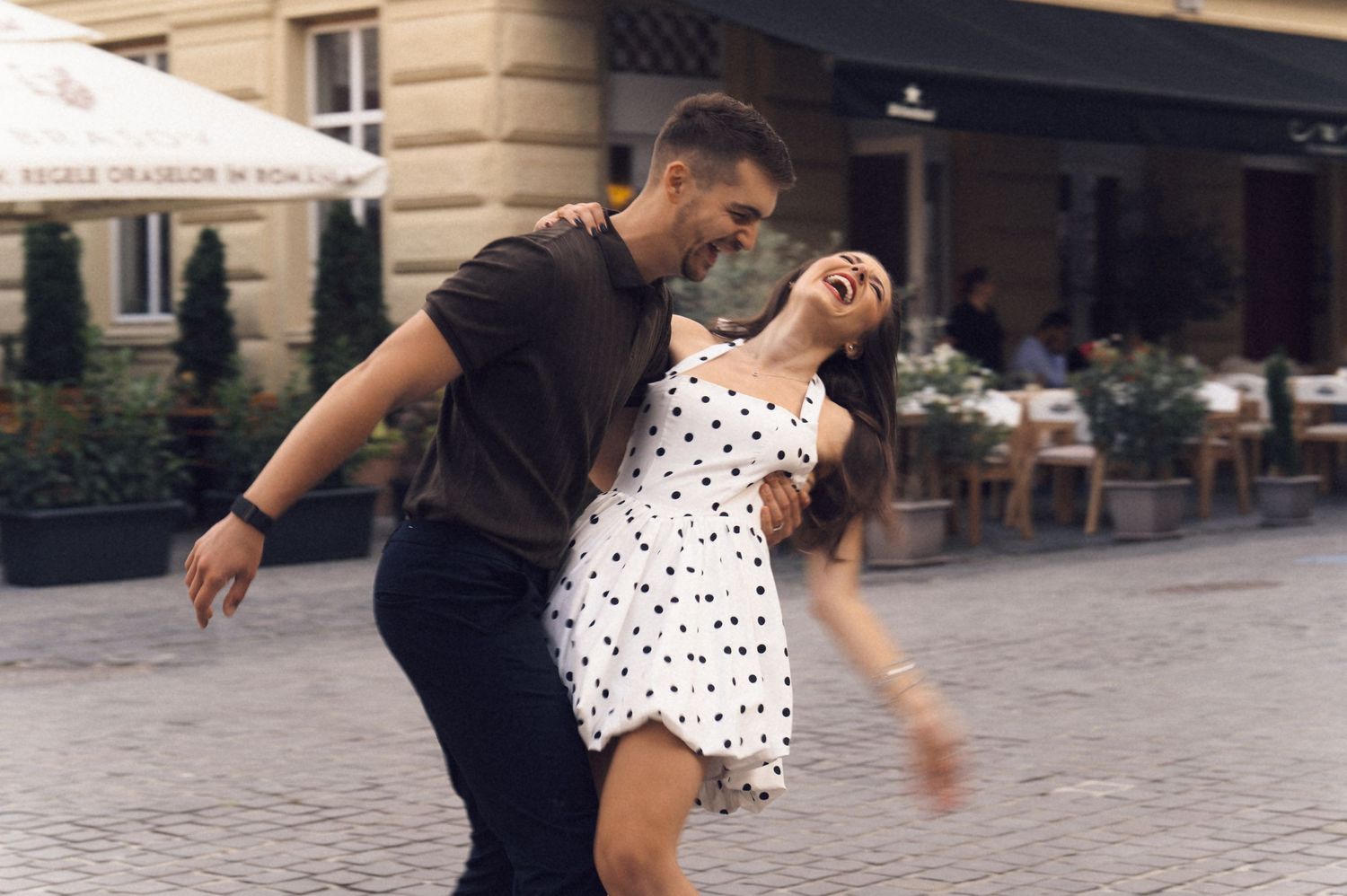 A couple dances joyfully together on a cobblestone street outside a cafe, with the woman wearing a polka dot dress.