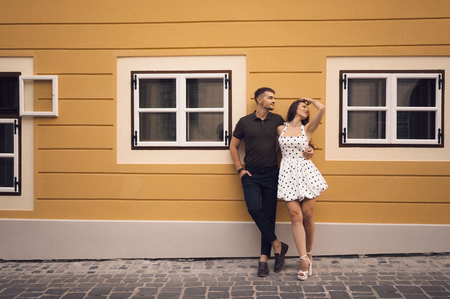 A couple in vintage-style clothing poses against a yellow building wall in a romantic photoshoot.