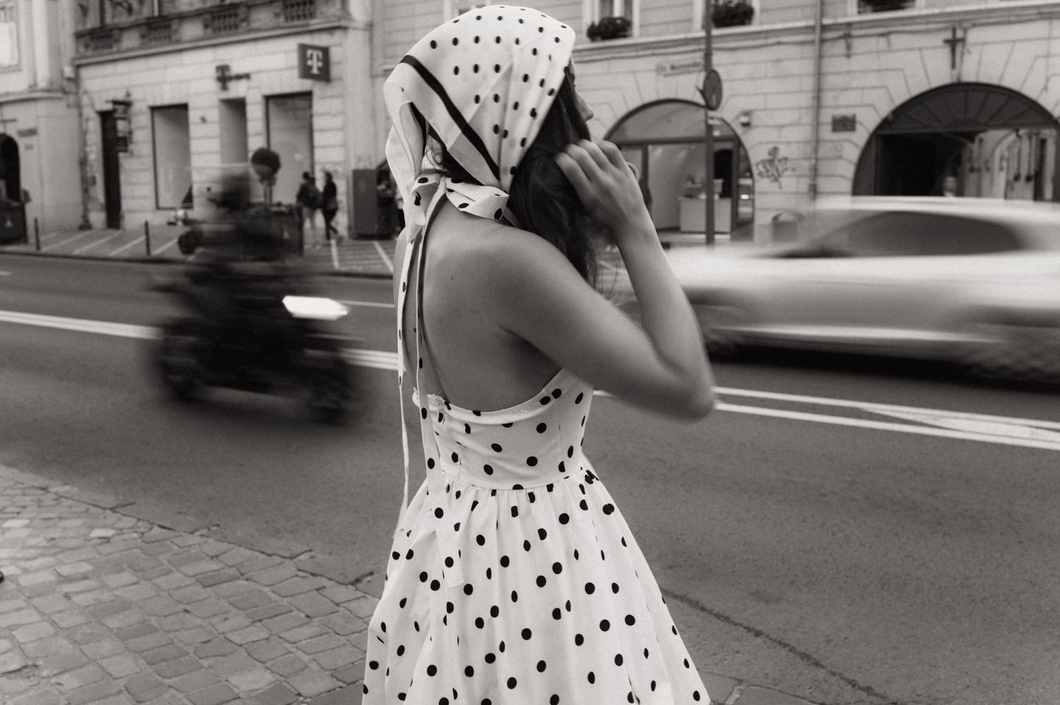 A person in a polka dot dress walks down a cobblestone street in a black and white photograph.