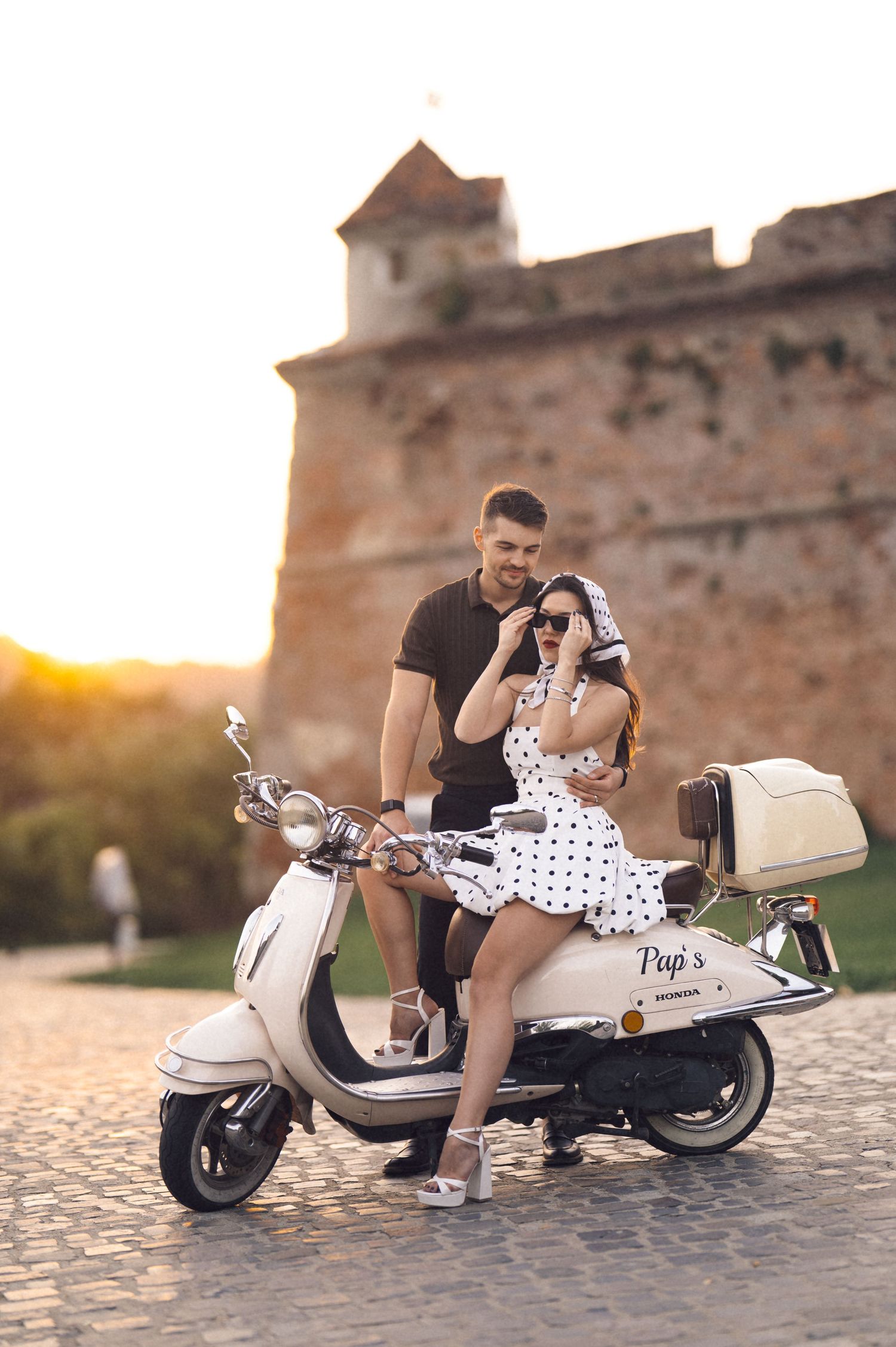 Romantic couple poses with a classic white Vespa scooter at sunset against a rustic stone wall backdrop.