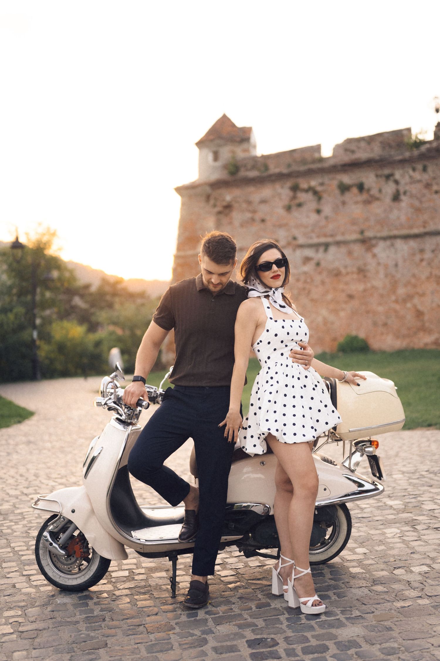 A couple sits together on a vintage white Vespa scooter against a rustic brick wall backdrop during sunset.