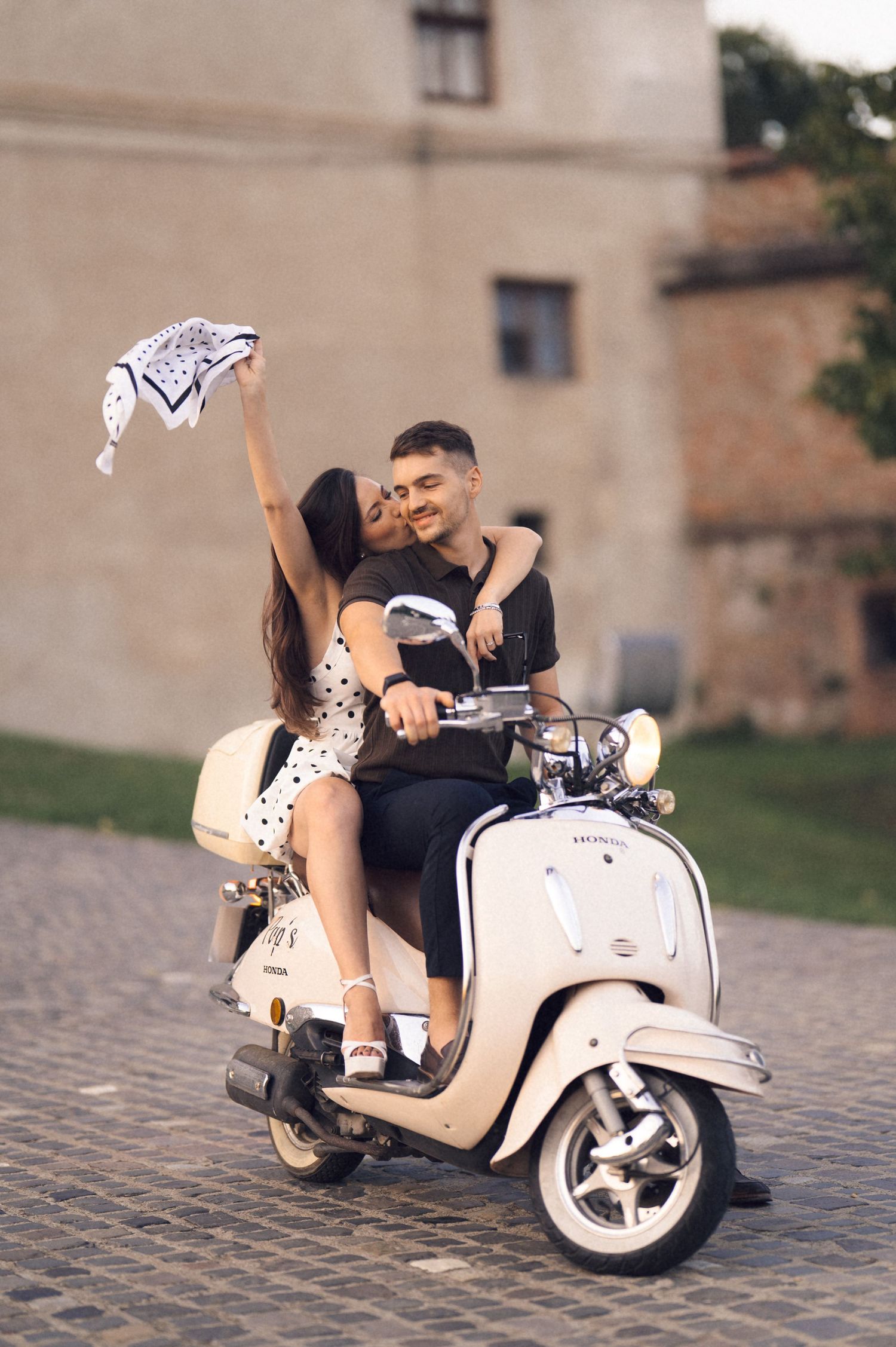 A couple shares a romantic moment on a vintage cream-colored Vespa scooter parked on cobblestone streets.