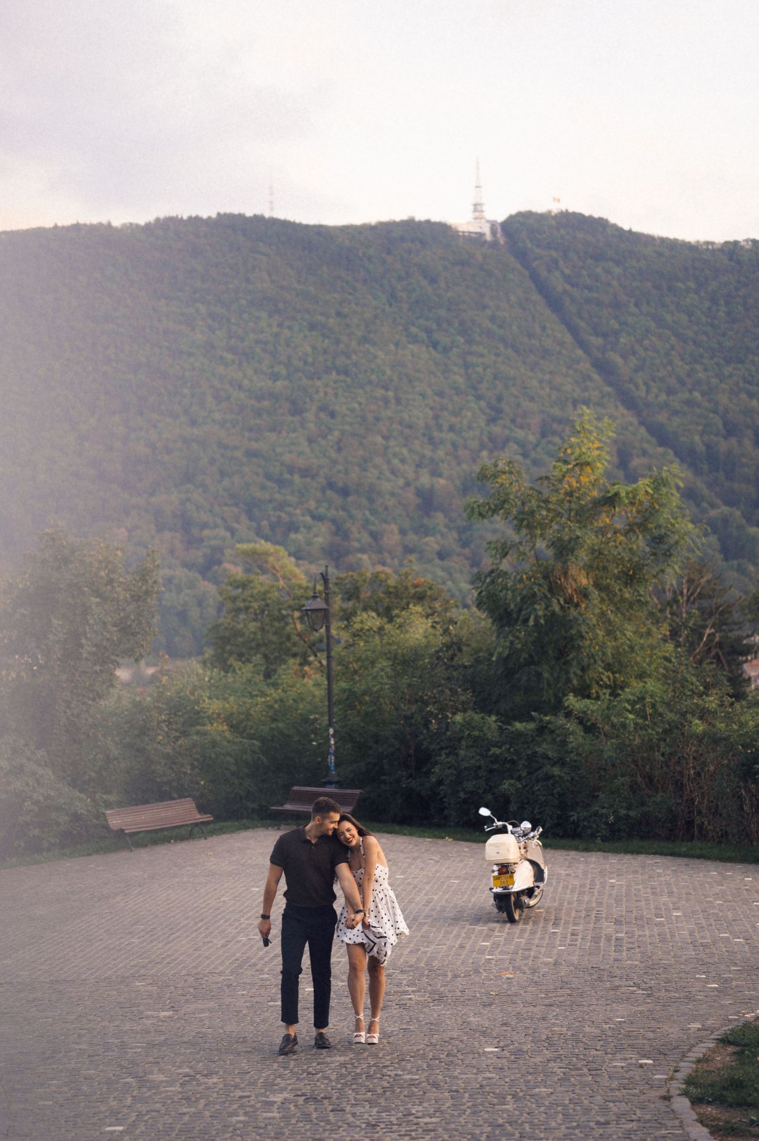 Two people and a small dog stand on a paved overlook with mountains in the background at sunset.