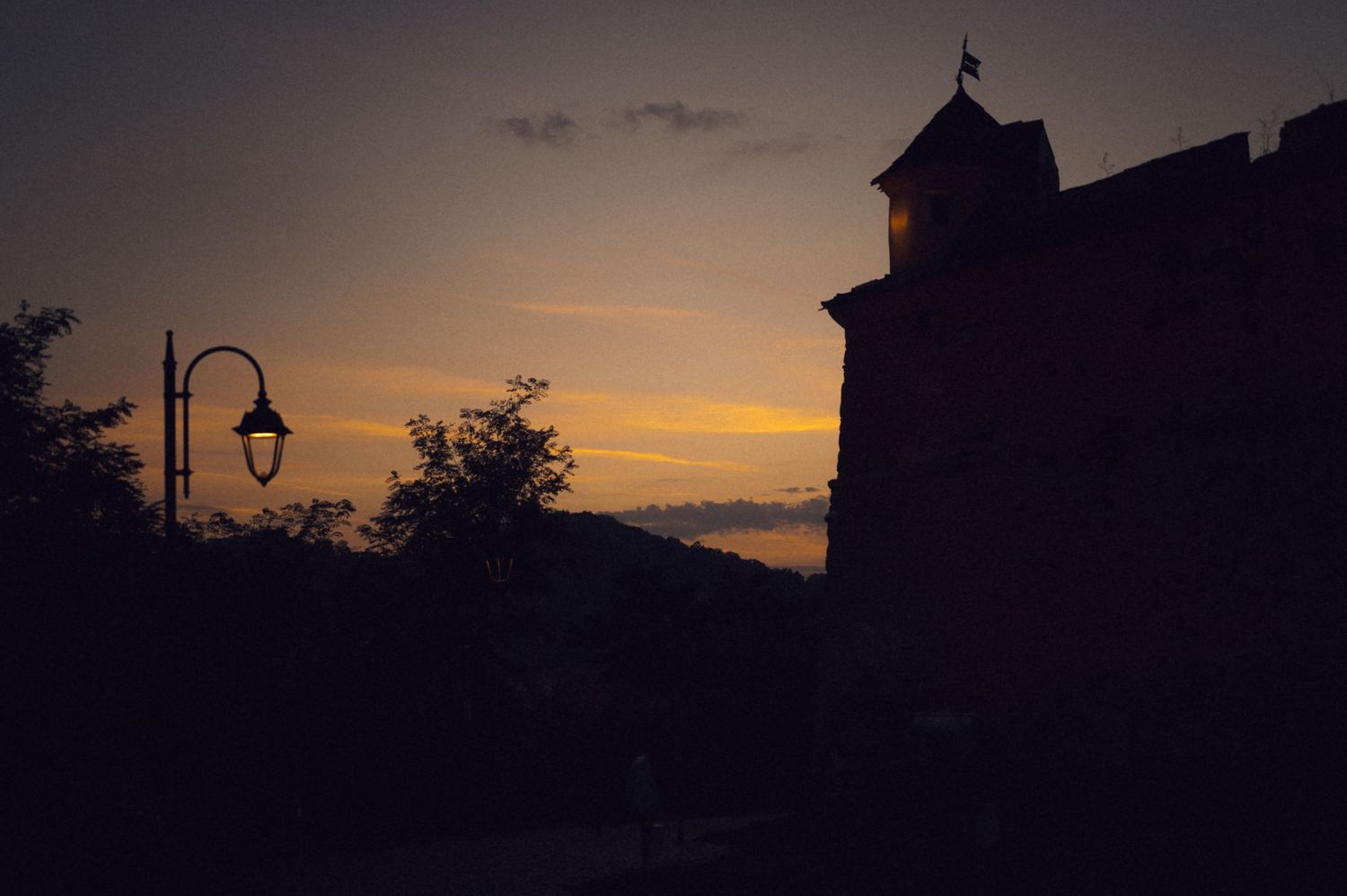 A dramatic sunset silhouettes trees and buildings against an orange and purple sky with a street lamp in view.