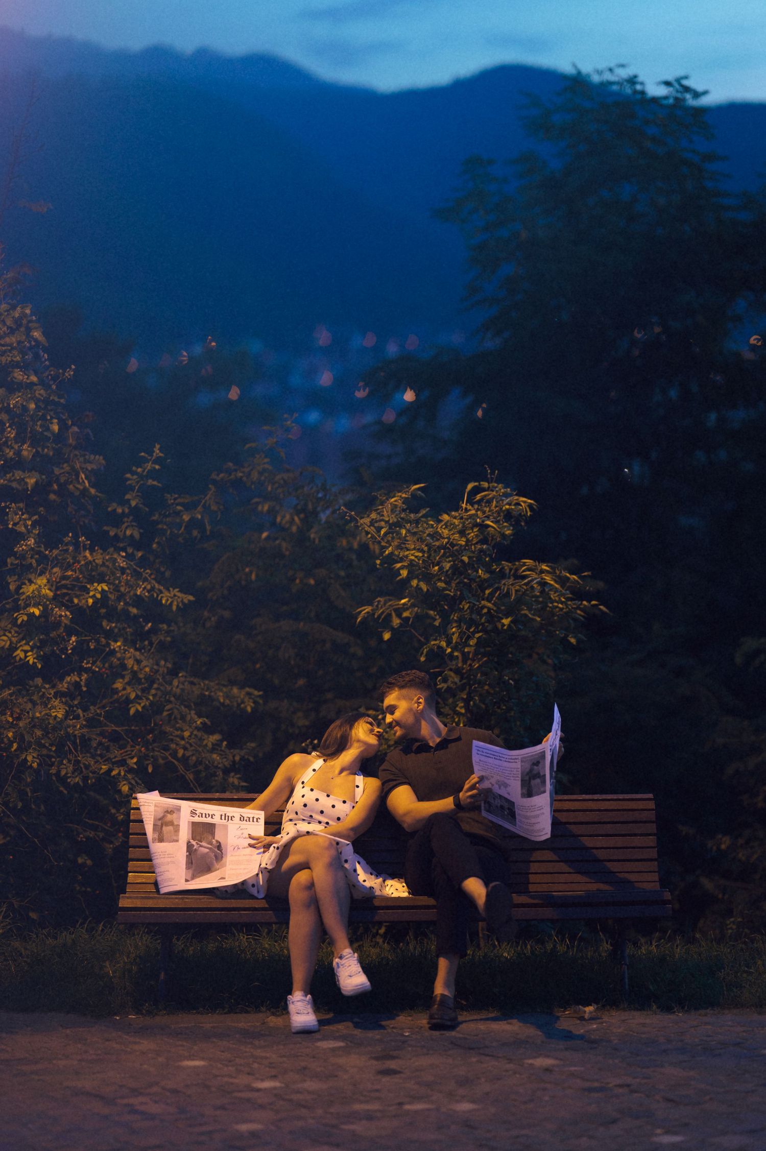 A couple sits on a park bench at dusk reading newspapers with mountains and city lights visible in the background.