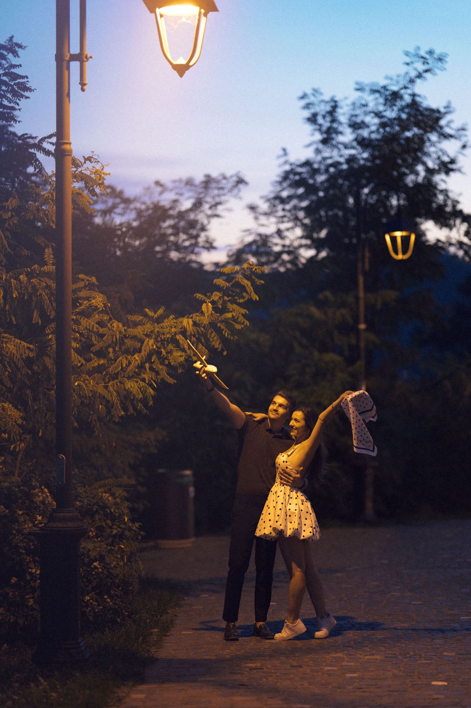 A couple poses playfully under glowing street lamps at dusk in a park setting.