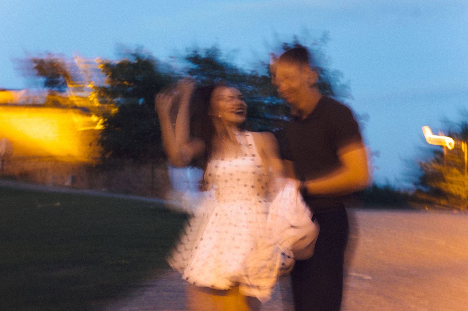 A blurred motion photo of a couple dancing outdoors at dusk with warm lighting in the background.