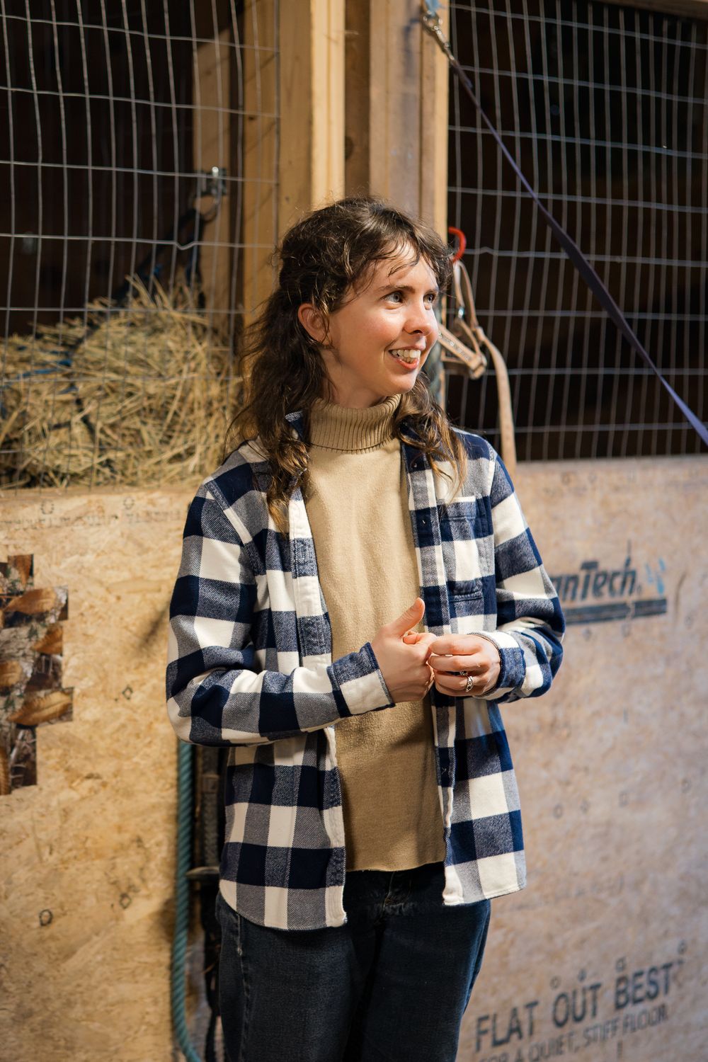 A person wearing a blue buffalo plaid flannel shirt over a beige sweater smiles while standing in a rustic barn setting.