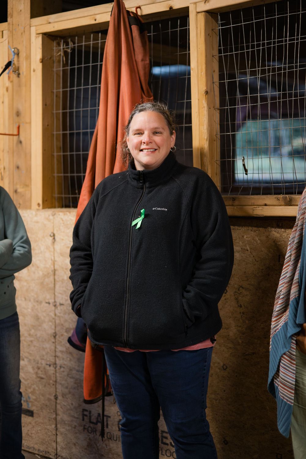 Person wearing a black jacket stands inside a rustic wooden barn stable with wire mesh windows.