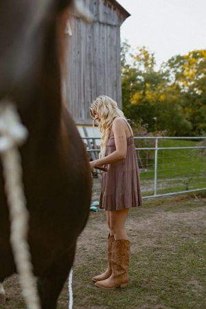 A person in a brown dress and cowboy boots stands near a rustic barn in a rural setting.