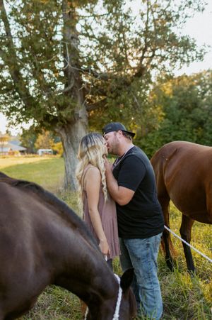 Two horses graze while people share a moment under a large tree at sunset.