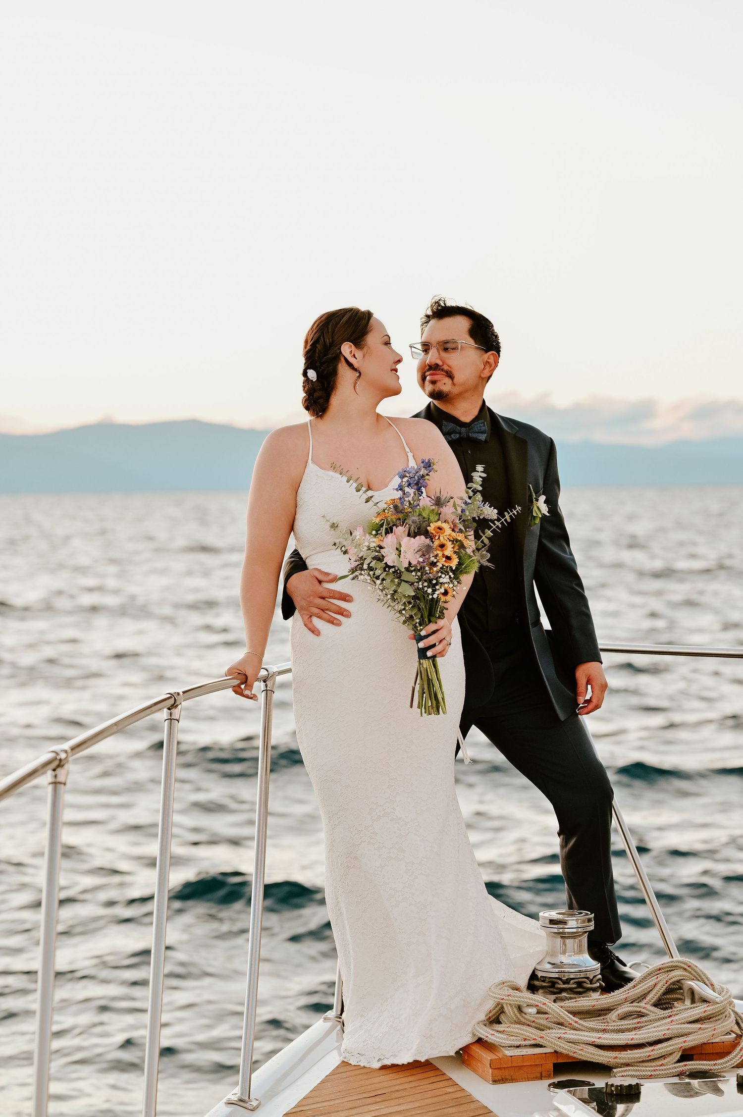 A romantic wedding couple shares intimate moments aboard a boat deck at sunset with mountains and water in the background.