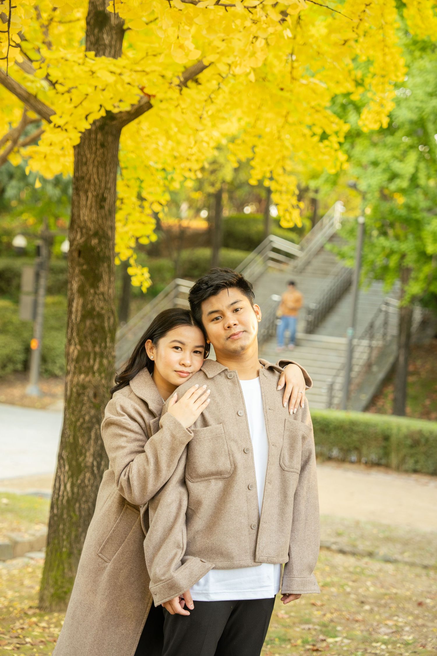 A couple poses together under vibrant yellow autumn trees in a park setting.