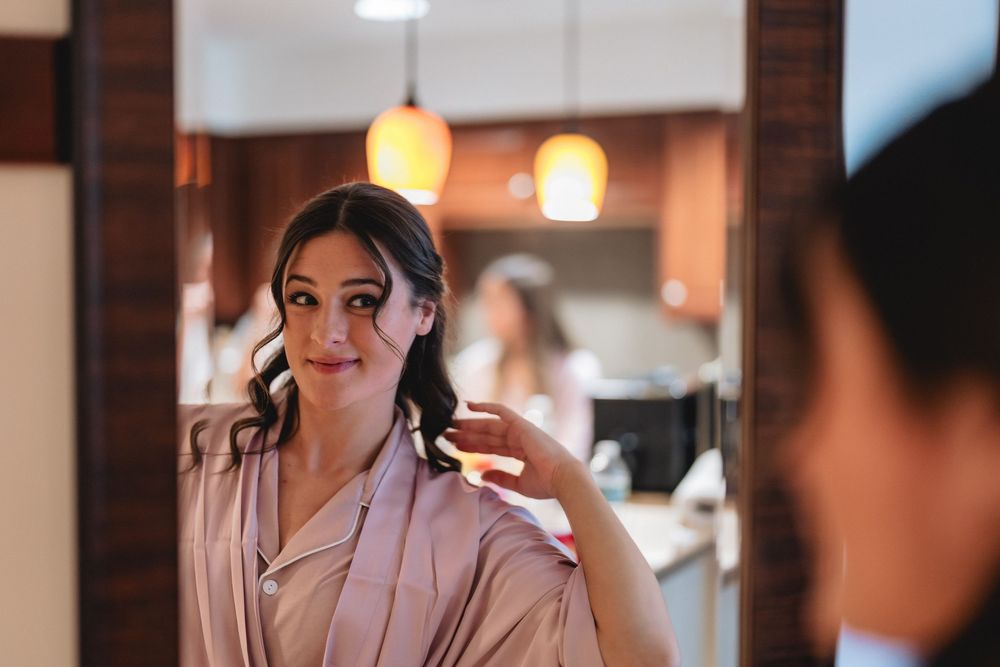 Woman wearing a beige robe stands pensively in front of a mirror with warm lighting in background.