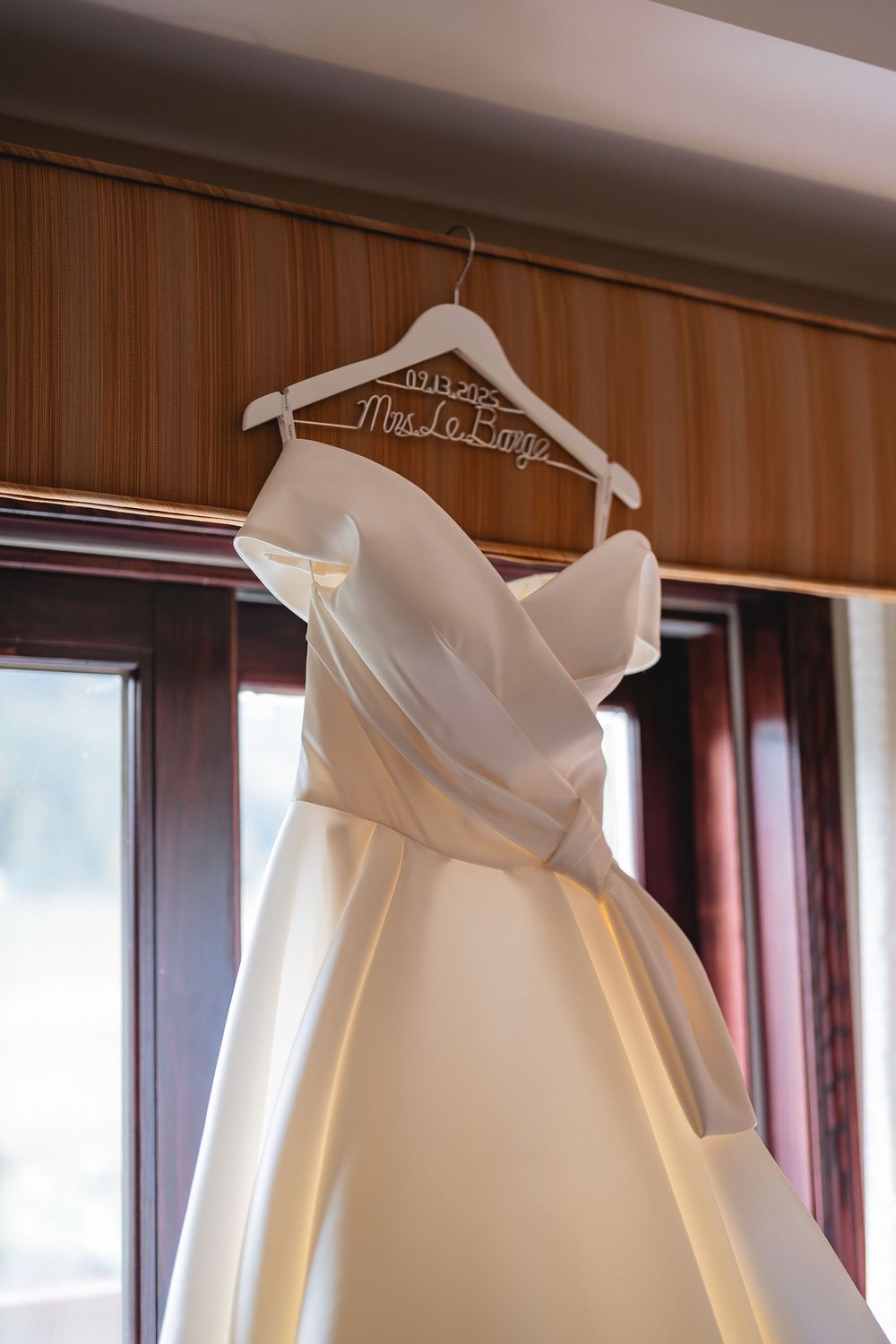 Row of identical white wedding dresses displayed on hangers against a wood paneled wall with window light.