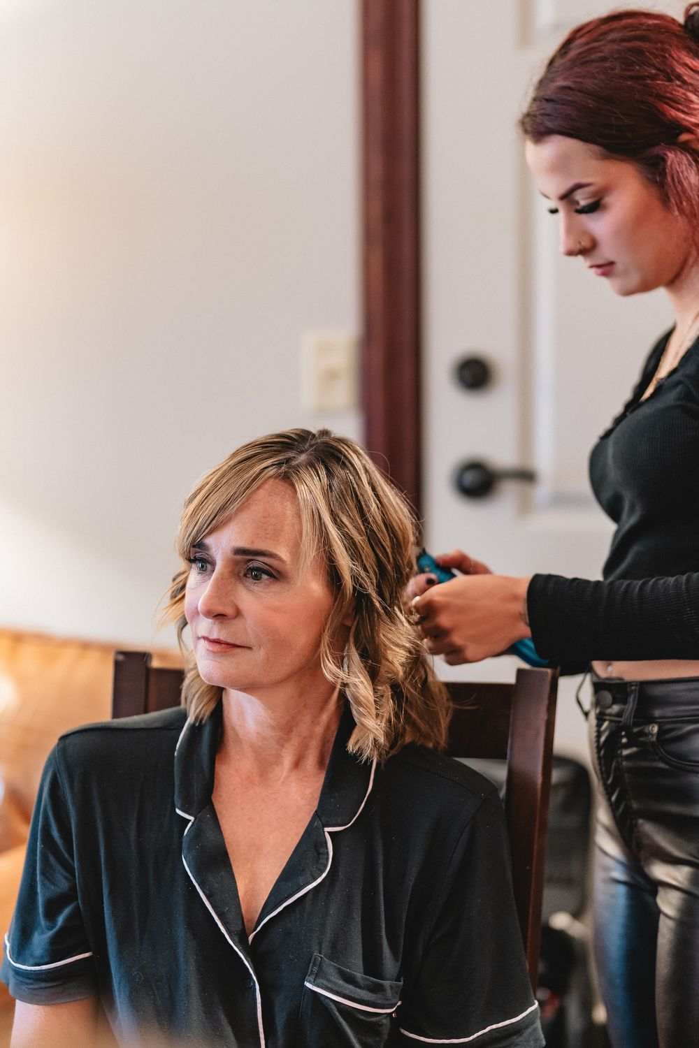 Woman in black sleepwear getting hair styled in salon chair while others work in background.