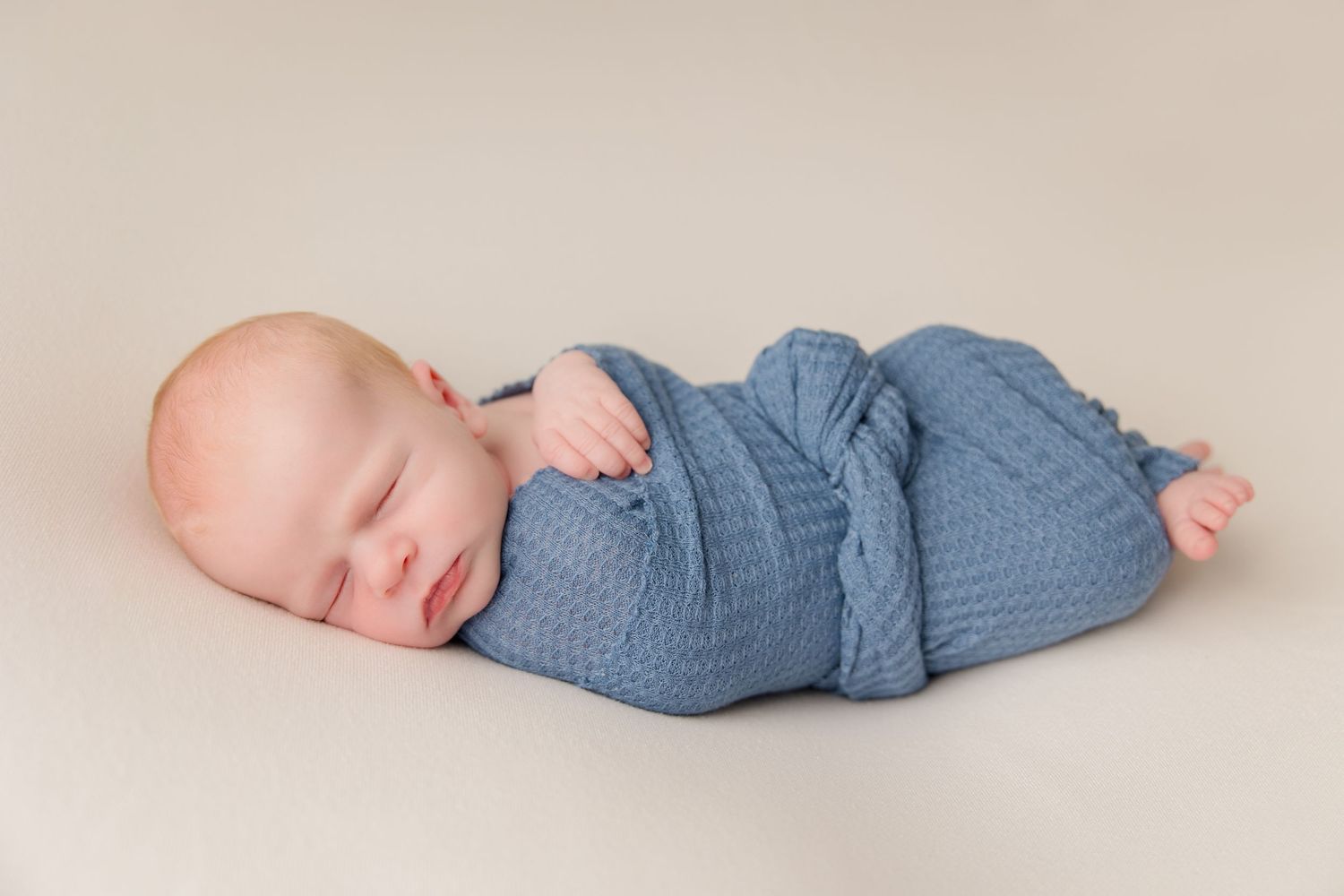 A baby wearing blue knit clothing rests peacefully on a beige background in minimalist style.