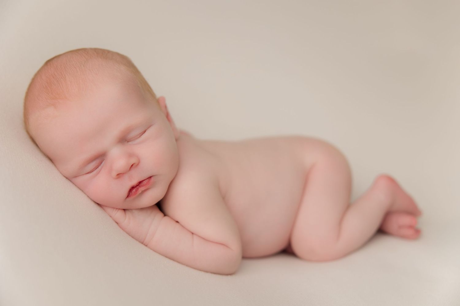 A sleeping newborn baby lies peacefully against a soft beige background in a minimalist portrait.