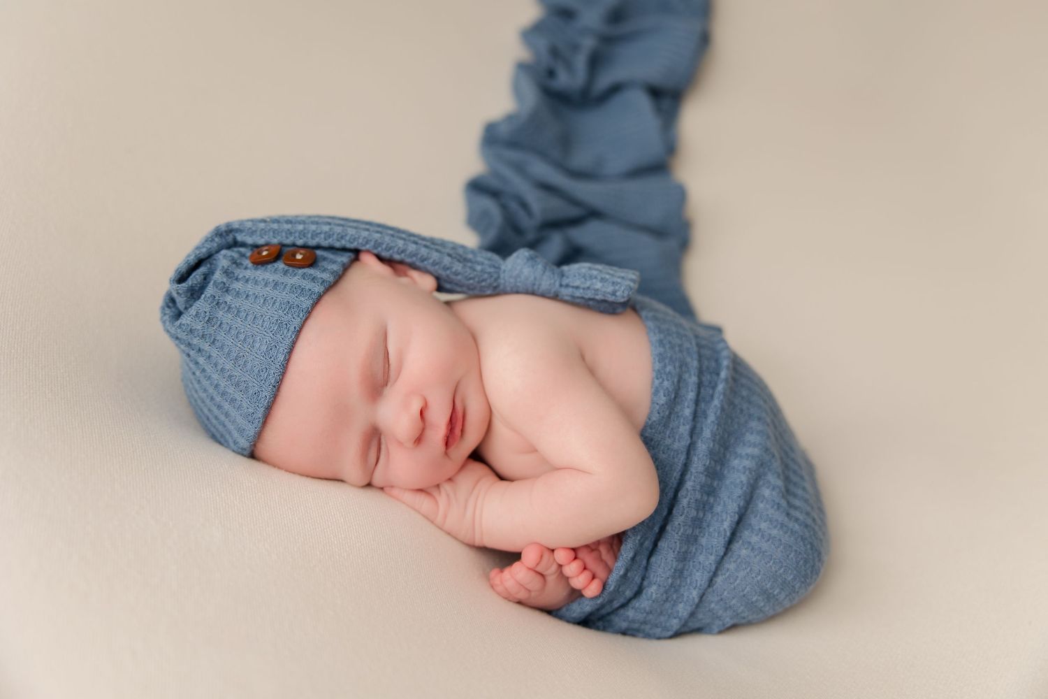 Newborn baby wearing denim outfit laying peacefully on cream colored backdrop.
