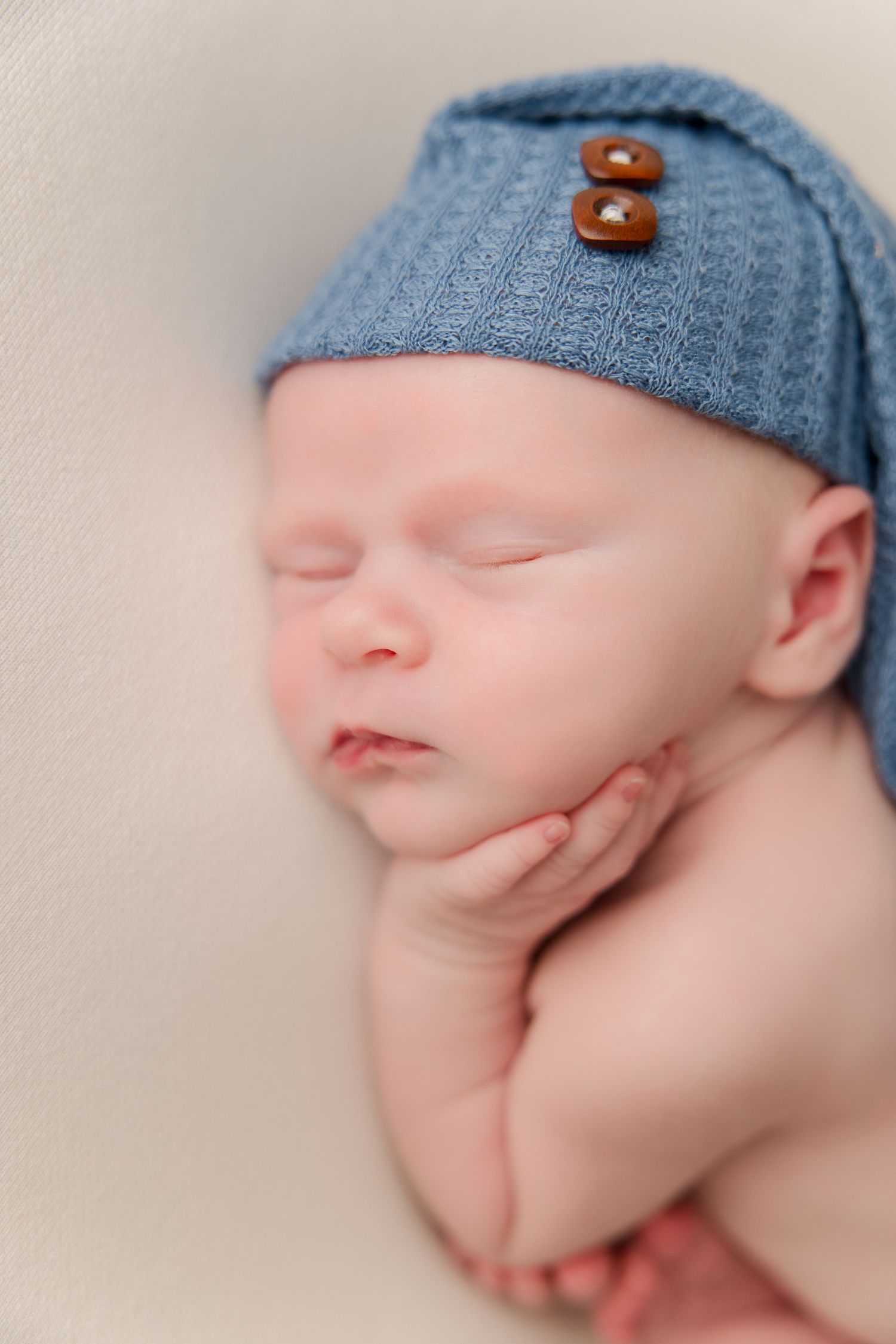 Sweet newborn sleeping peacefully in blue knit cap with decorative wooden buttons on light neutral background.