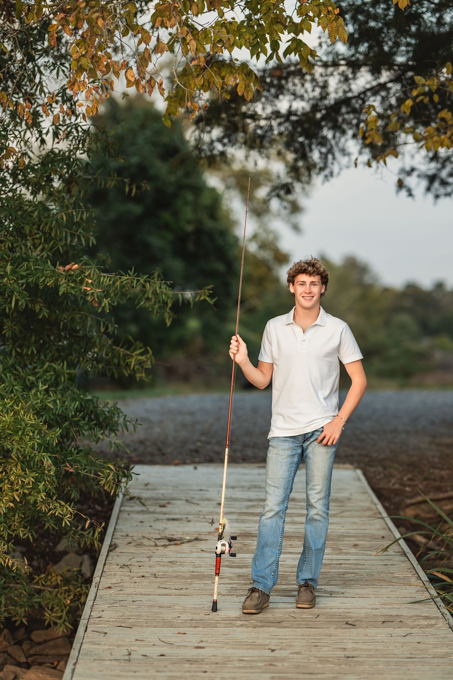 Senior boy in casual attire standing on wooden dock with fishing pole during autumn afternoon.
