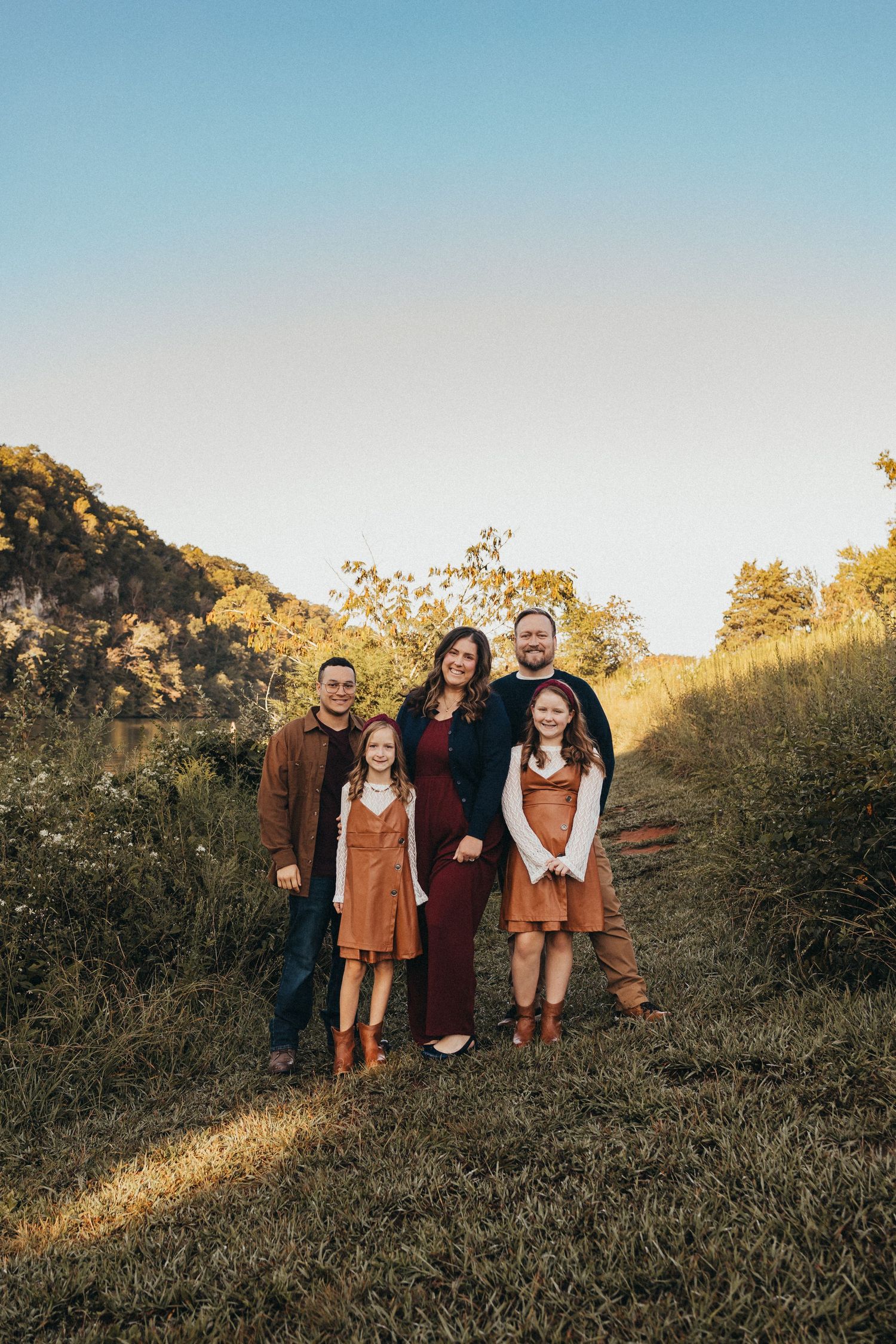 Family of five smiling together on a sunlit grassy trail with autumn trees and a hillside in the background.