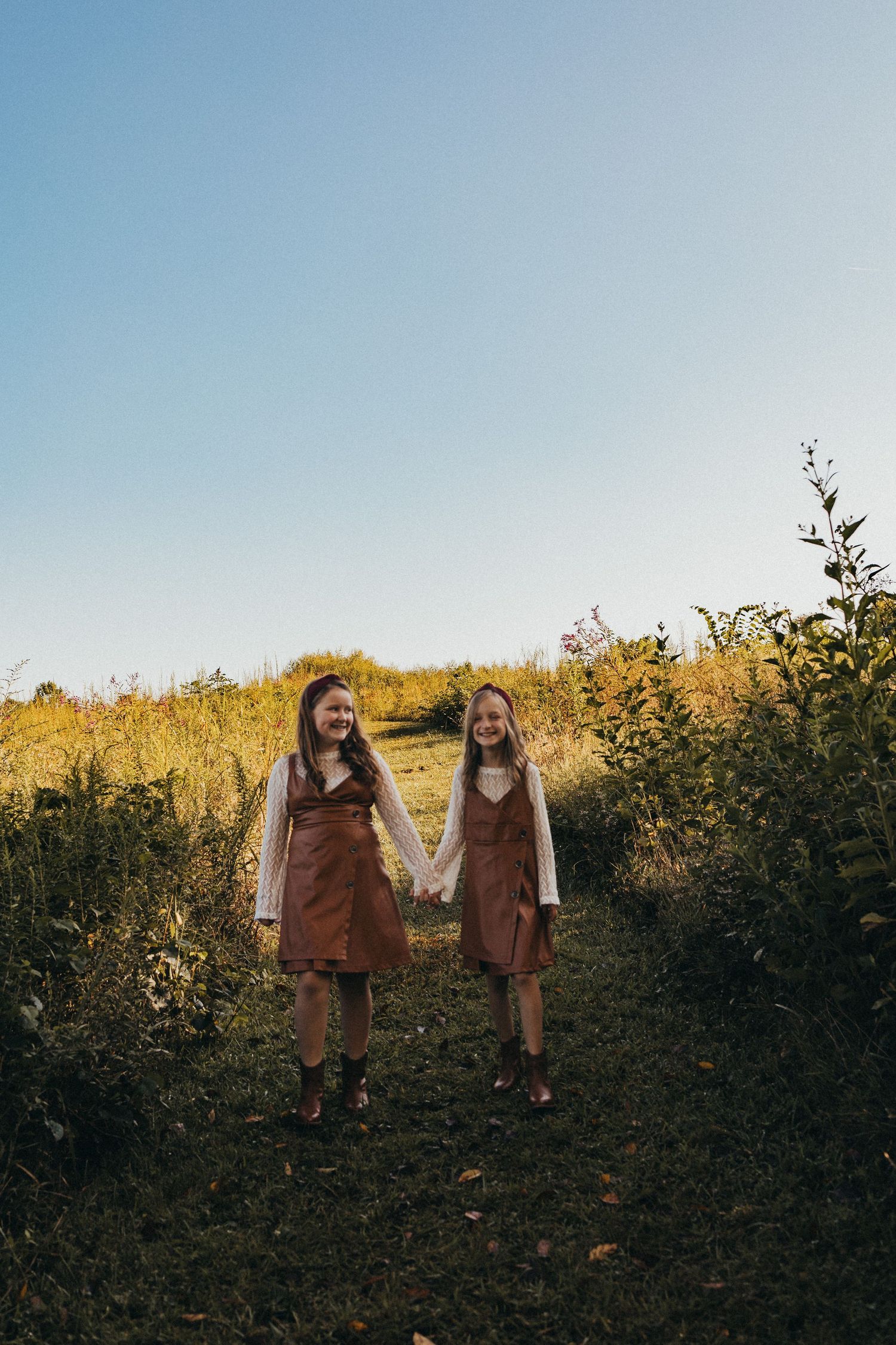Two girls in matching brown dresses hold hands while walking through a sunny field.