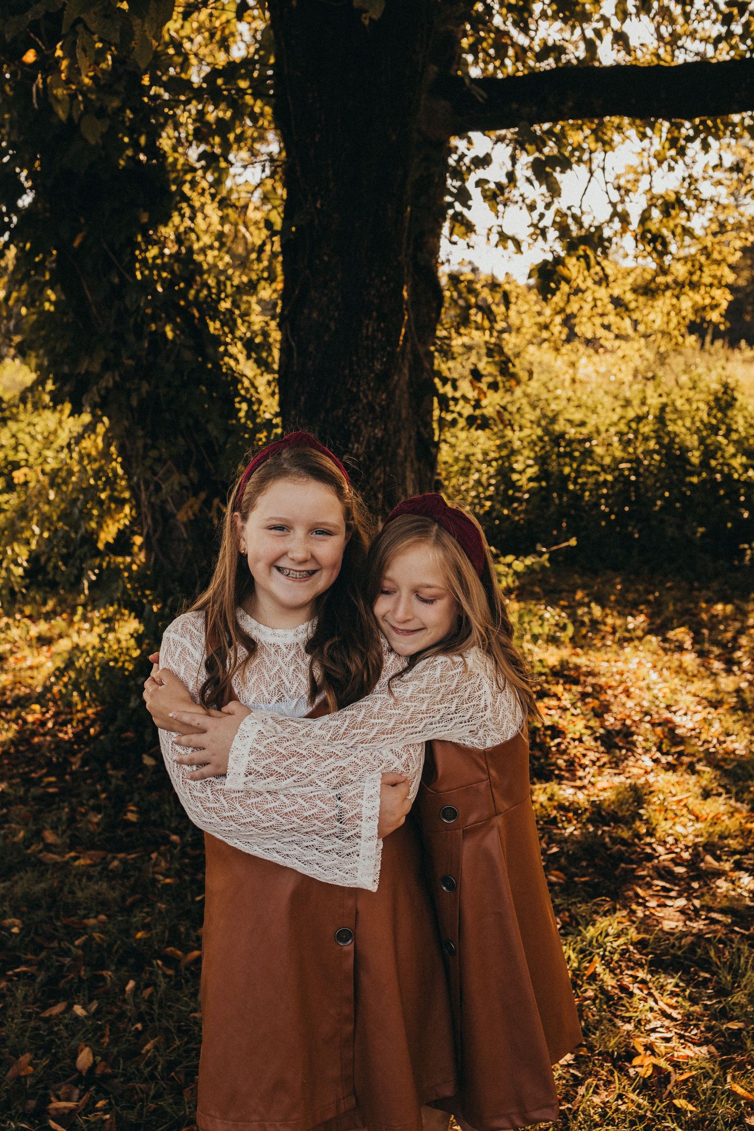 Two smiling girls in brown dresses embrace warmly in front of a sunlit autumn tree.