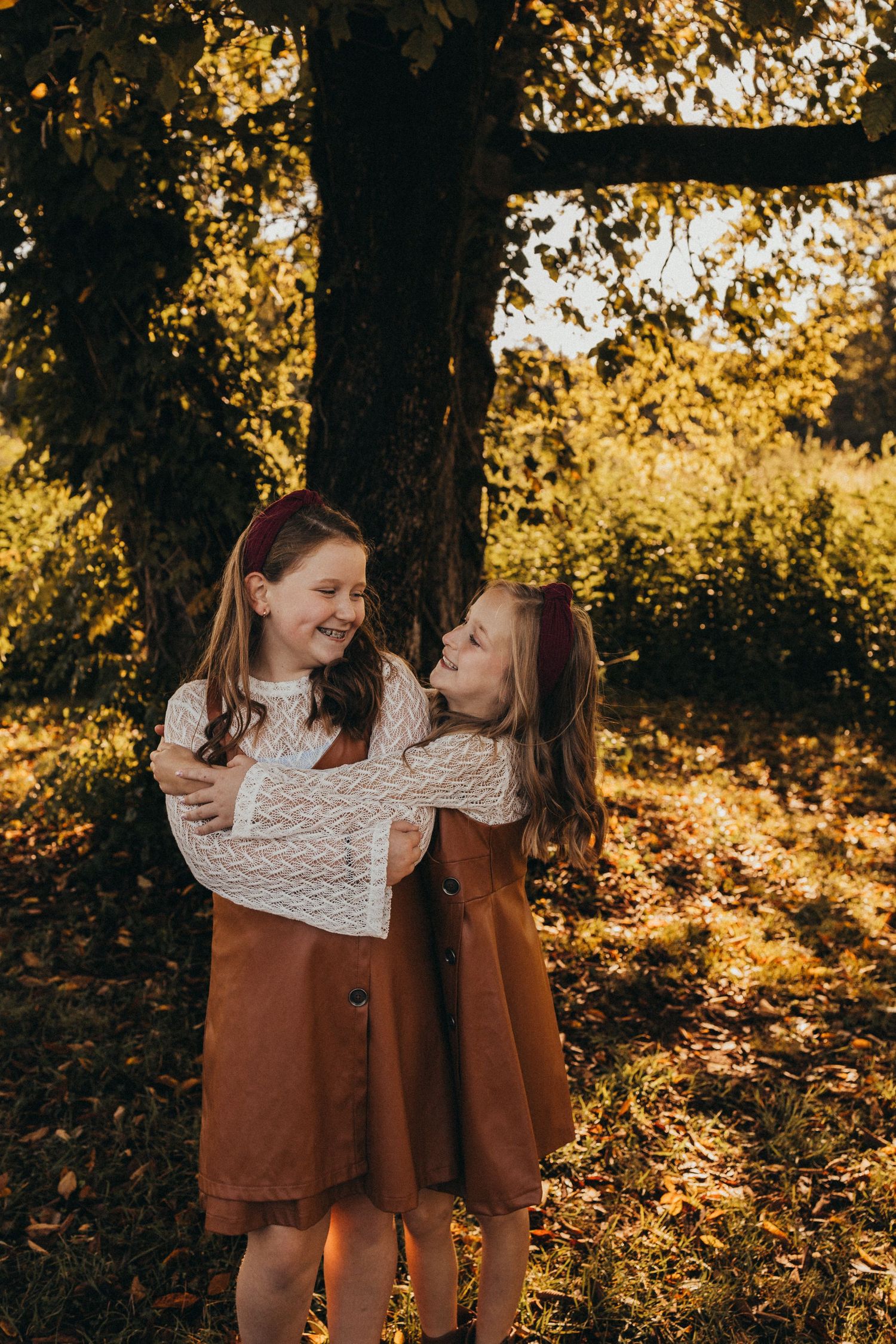 Two girls in matching brown dresses laugh and hug each other beside a golden autumn tree.