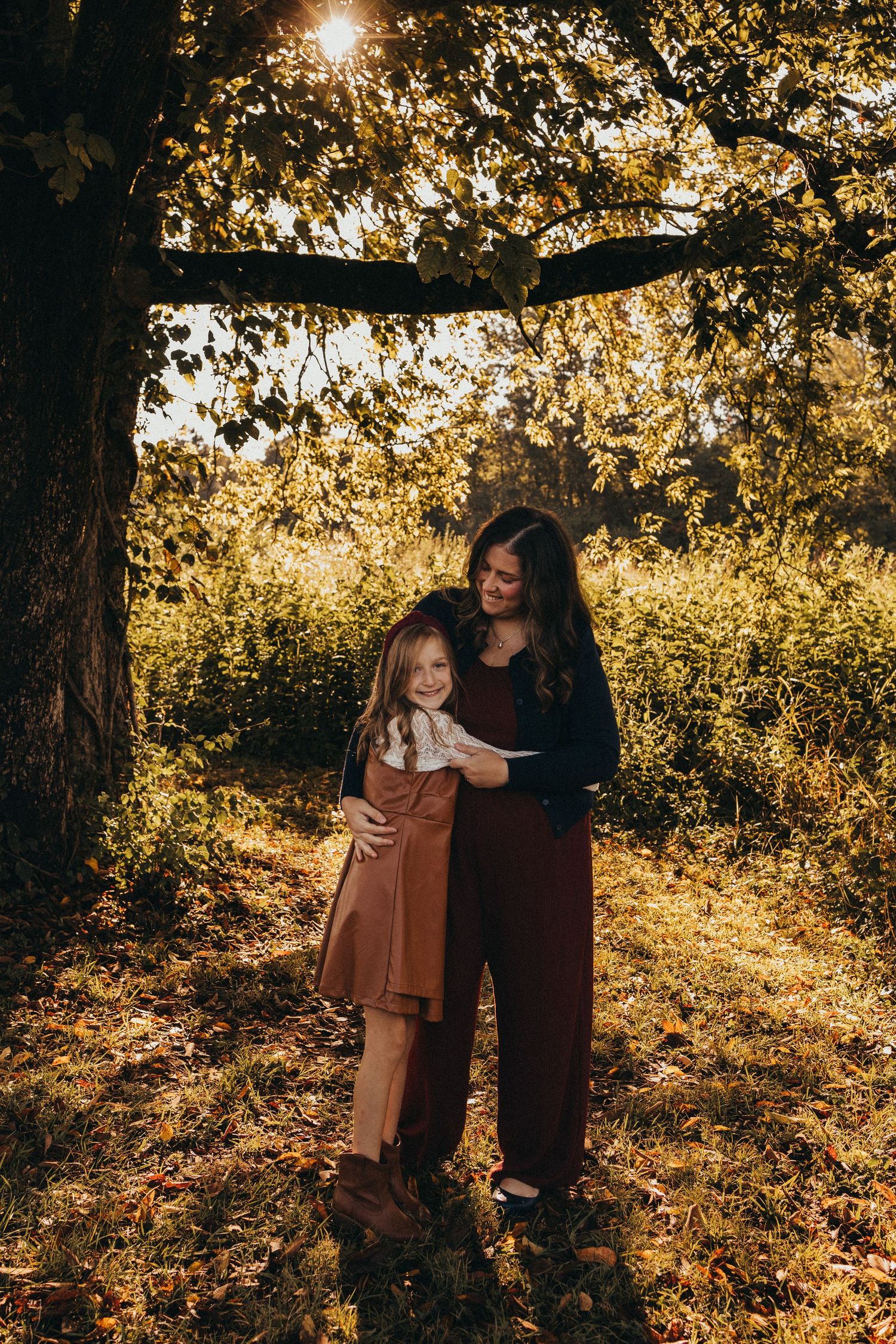 A mother in dark clothing tenderly embraces her young daughter in a sunlit autumn park.