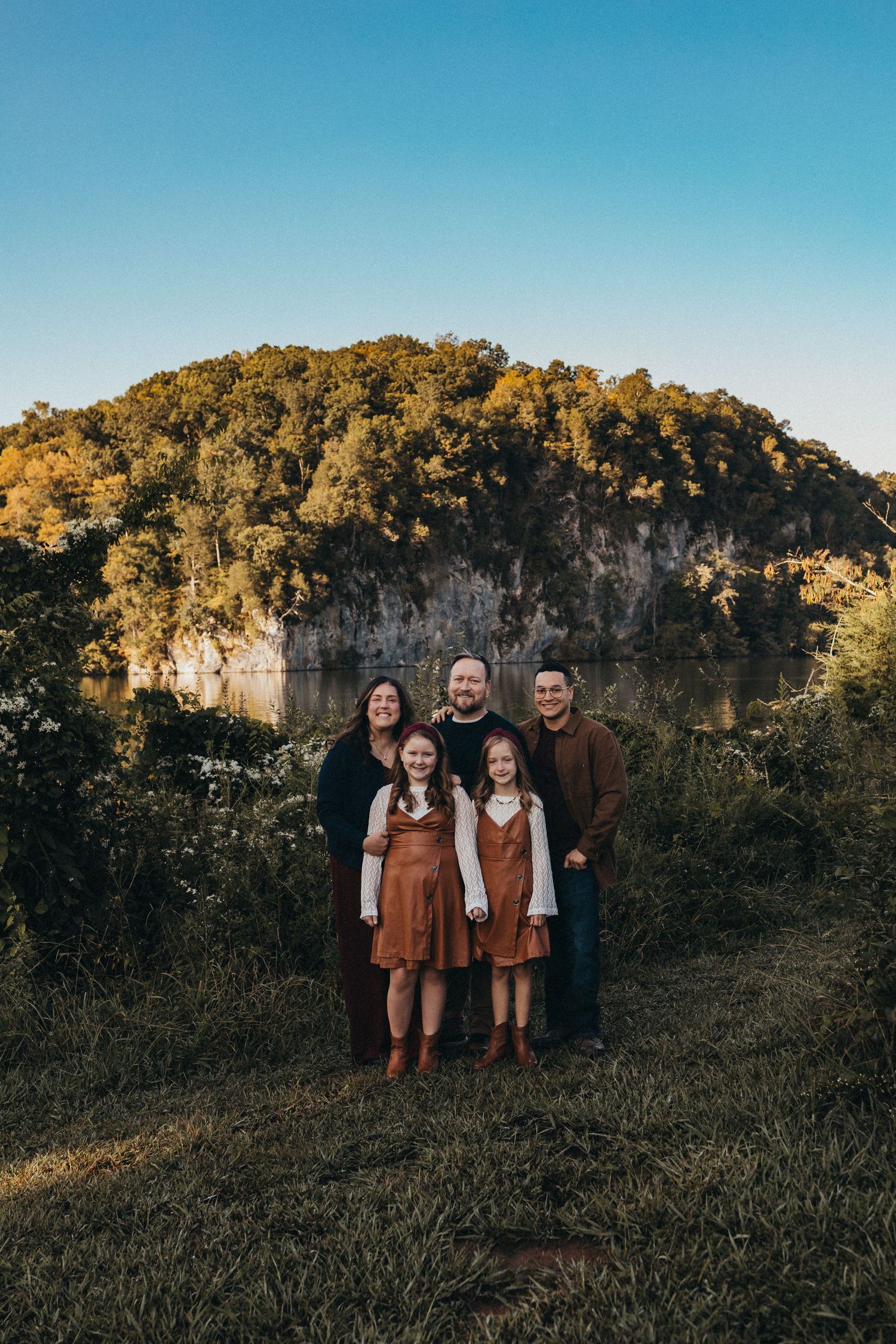 Family of five standing together in front of a scenic rocky bluff and lake surrounded by fall foliage.