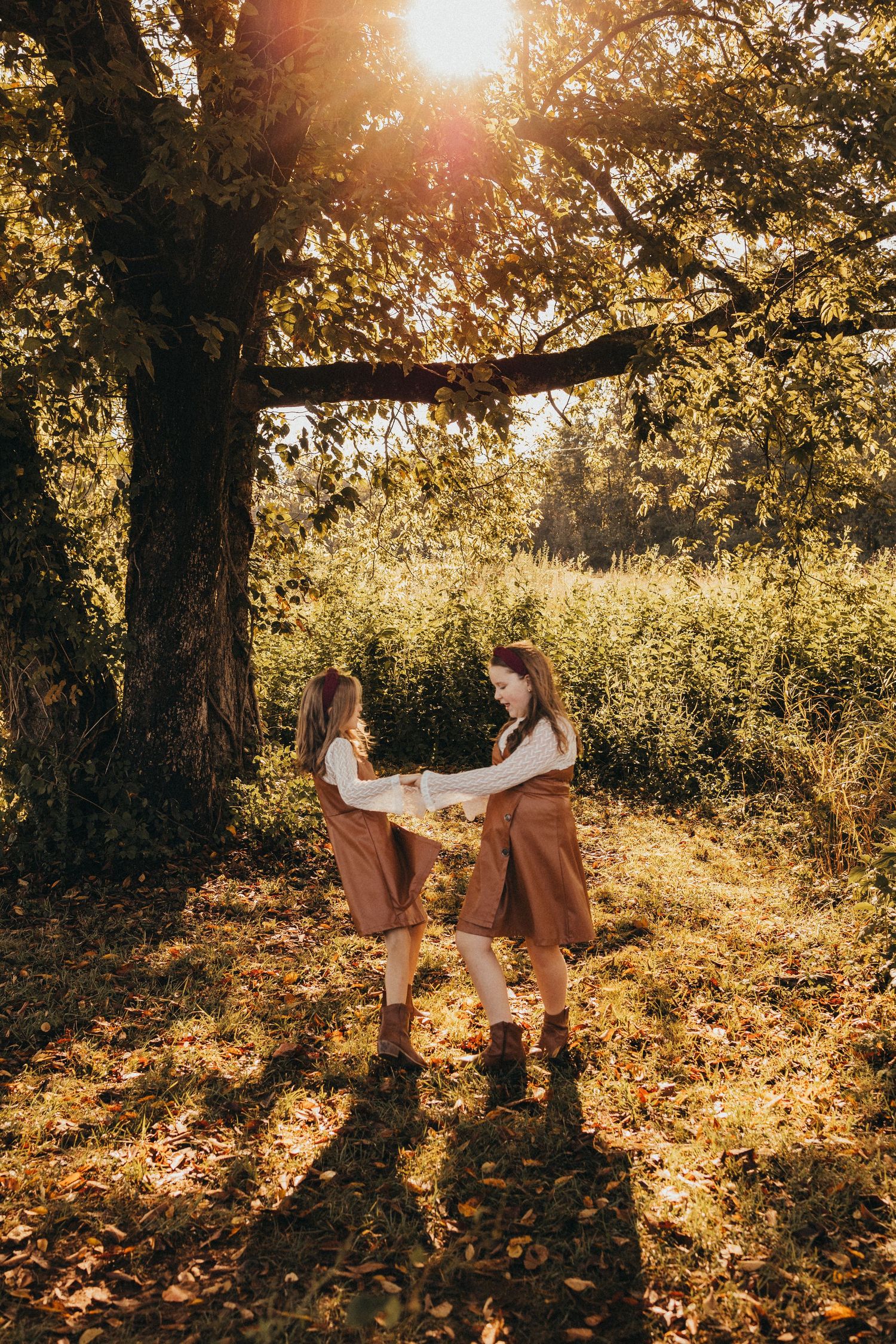 Two girls in matching brown dresses hold hands and spin together under a large sunlit tree.